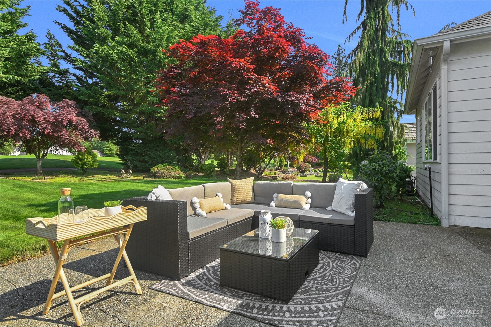 Furnished patio with a wicker sectional overlooks a green lawn and mature trees.