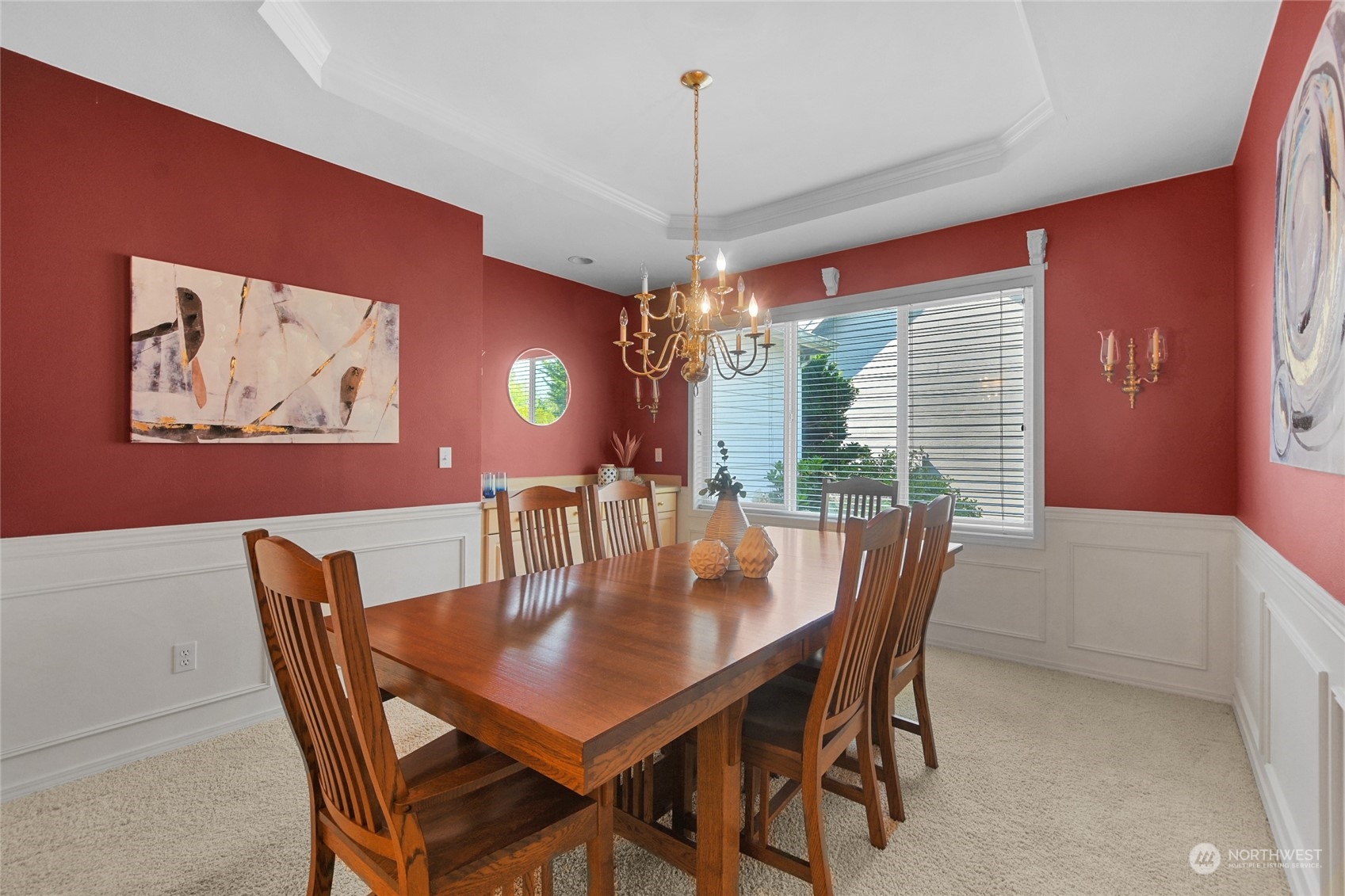 Dining room with a wooden table, chandelier, red walls, white wainscoting, and a wide window.