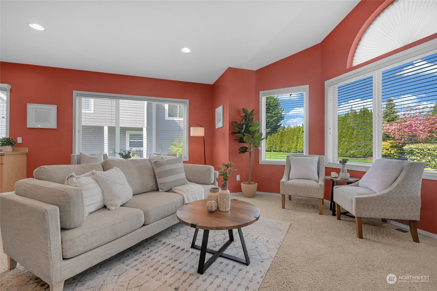 Furnished living room with coral walls, a gray sofa, and large windows overlooking a landscaped yard.
