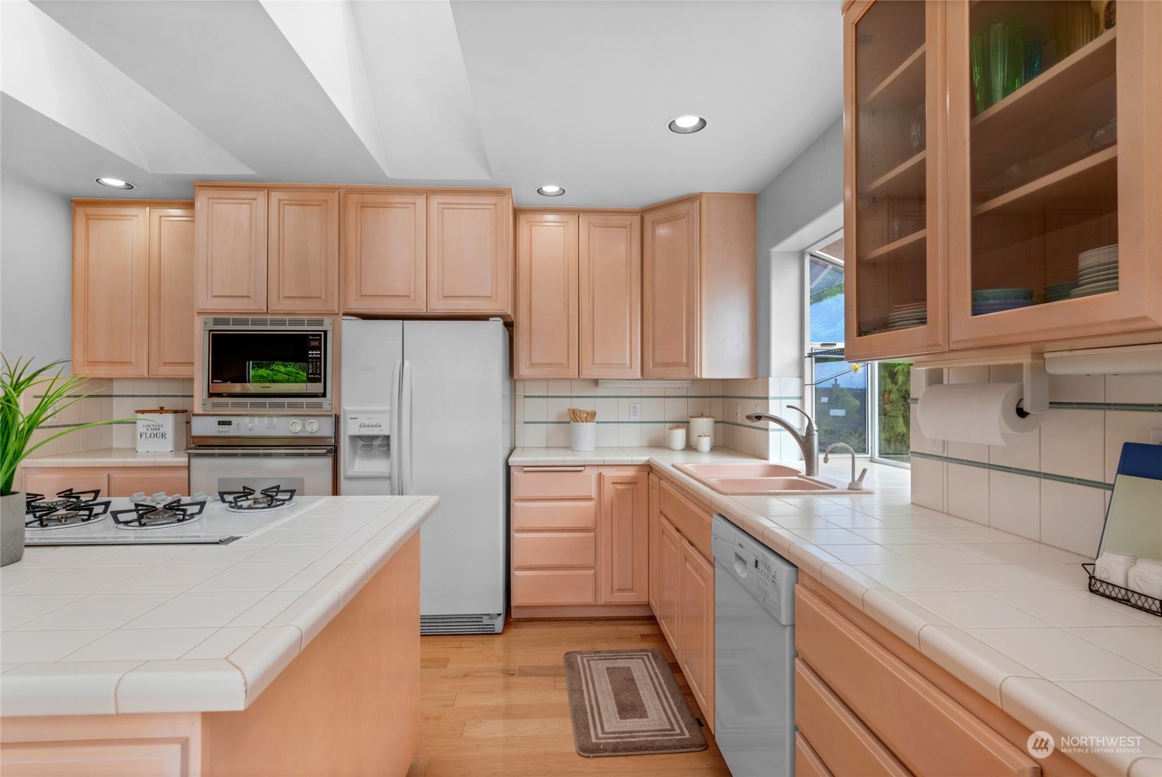 Kitchen with peach-toned cabinets, white tile counters, a central cooktop, and a double sink by the window.