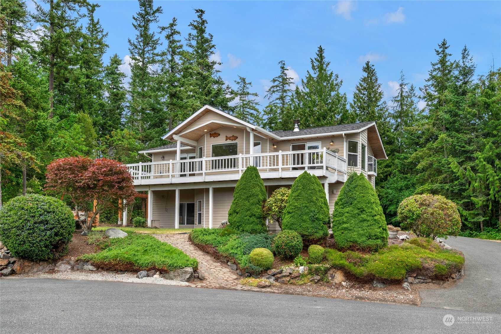 Beige two-story home with a broad white-railed deck, landscaped garden, and wooded surroundings.