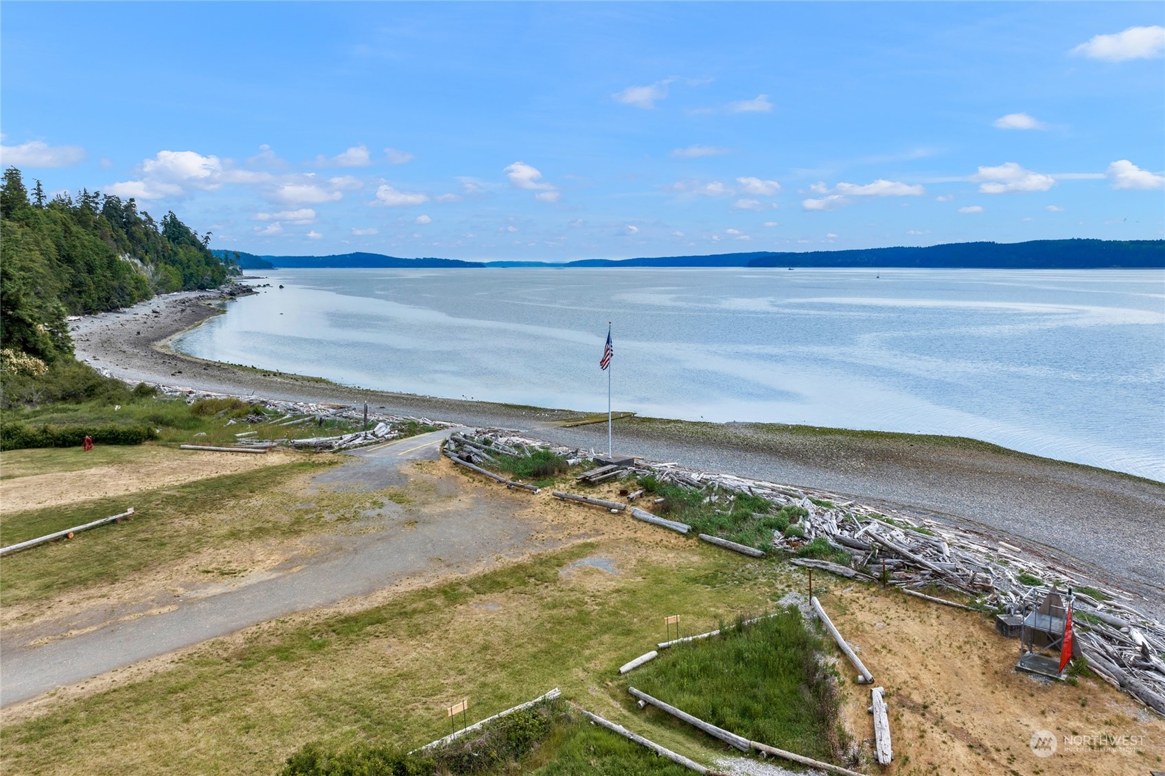 Waterfront shoreline with calm water, driftwood-covered beach, grassy foreground, and an American flag.