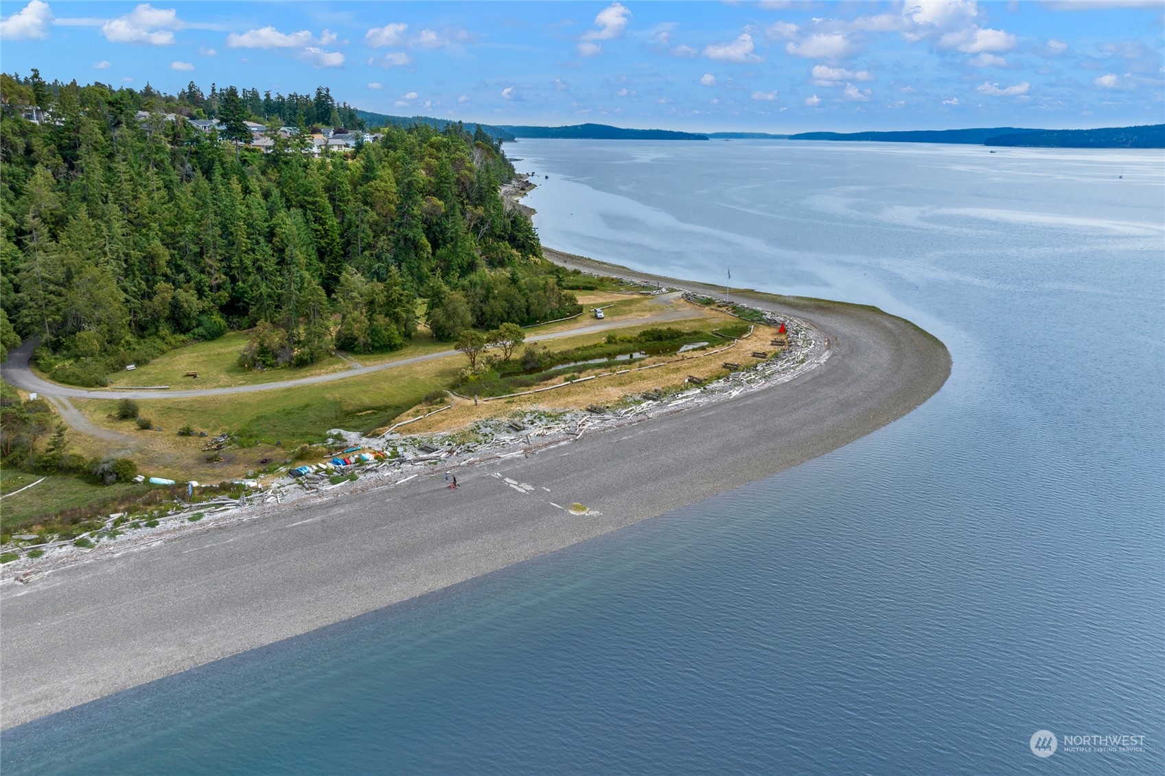 Aerial shoreline with a curved pebble beach, wooded land, and calm water.