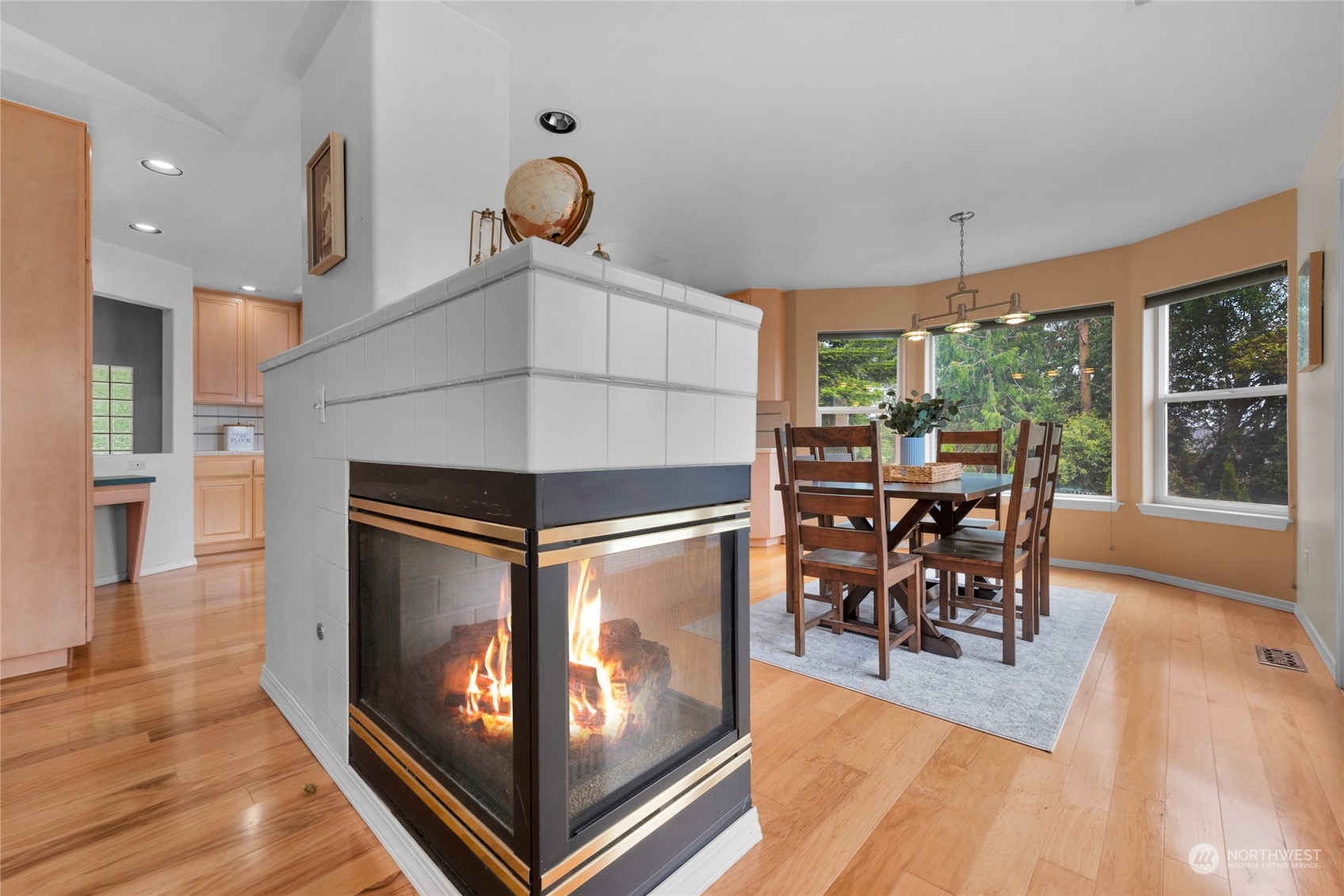 Dining area with a lit two-sided fireplace, hardwood floors, and large windows overlooking trees.