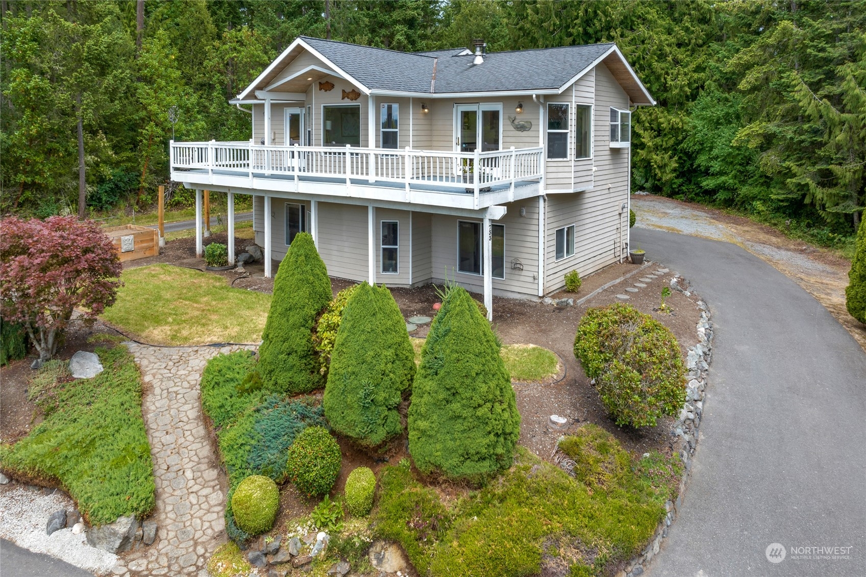 Two-story beige house with a white-railed deck, landscaped garden, and stone walkway among evergreen trees.