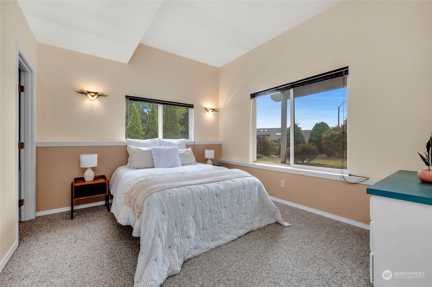 Staged bedroom with a white bed beneath a high ceiling and wide windows overlooking the yard.
