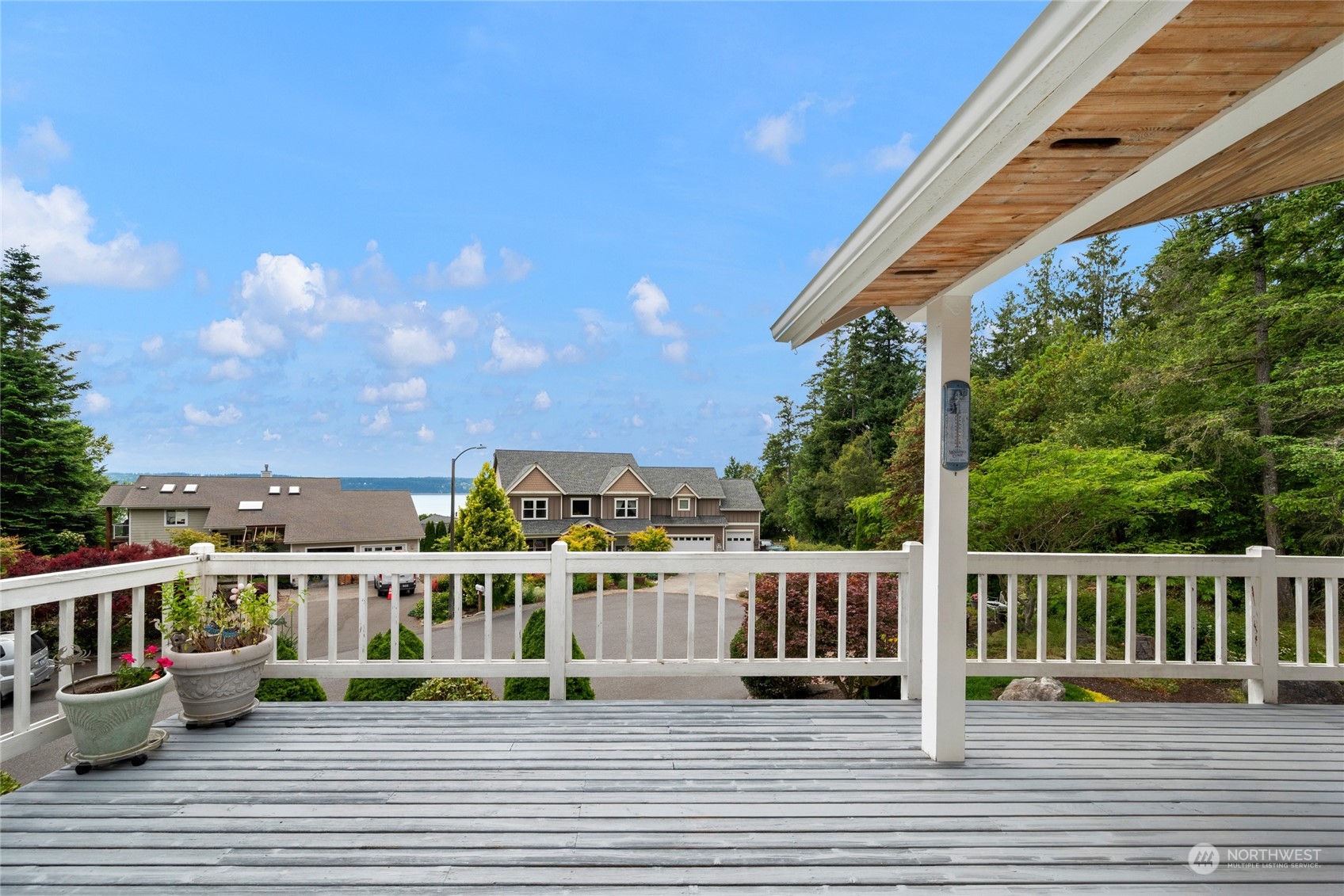 Deck with white railings and potted flowers overlooking homes, trees, and a glimpse of water.