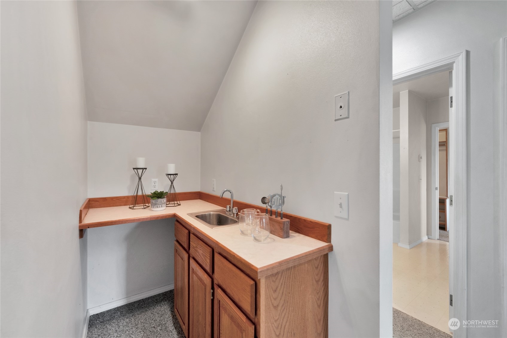 Compact sink nook with warm wood cabinets, a light countertop, and staged glasses beneath a sloped ceiling.