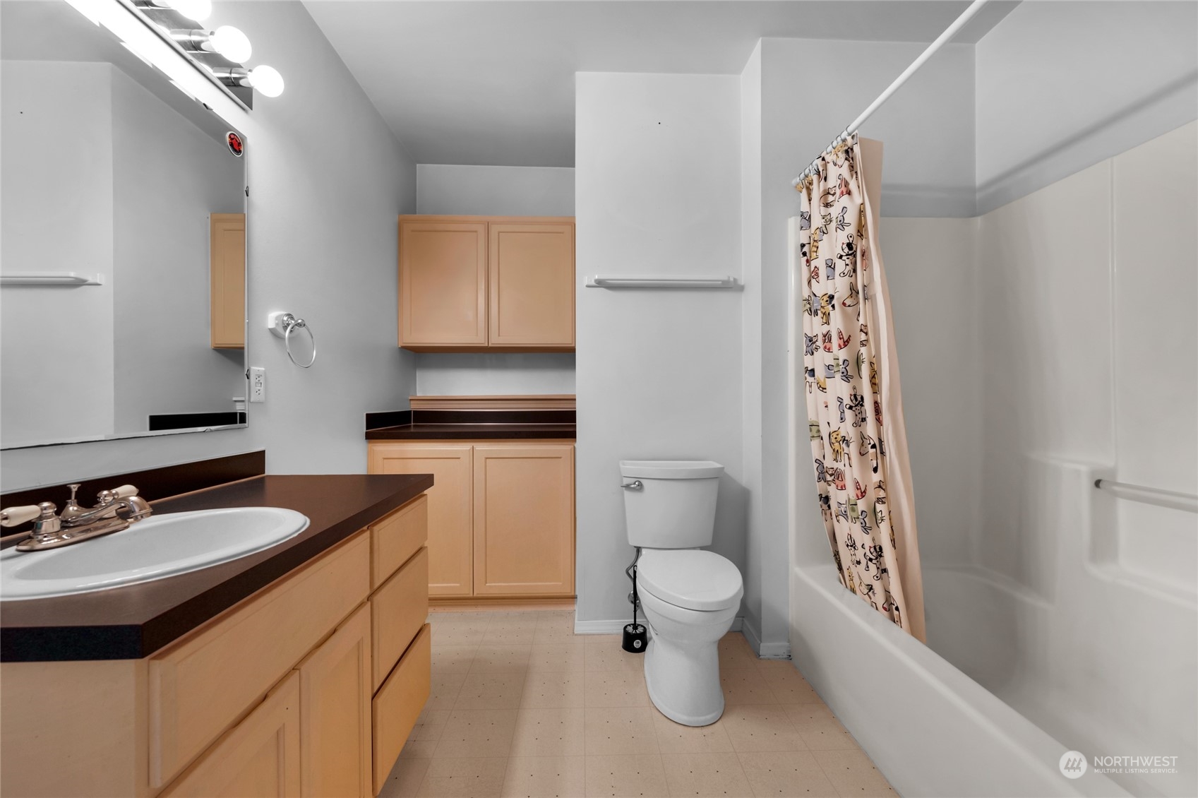Bathroom with a dark vanity countertop, light wood cabinets, toilet, and patterned shower curtain.