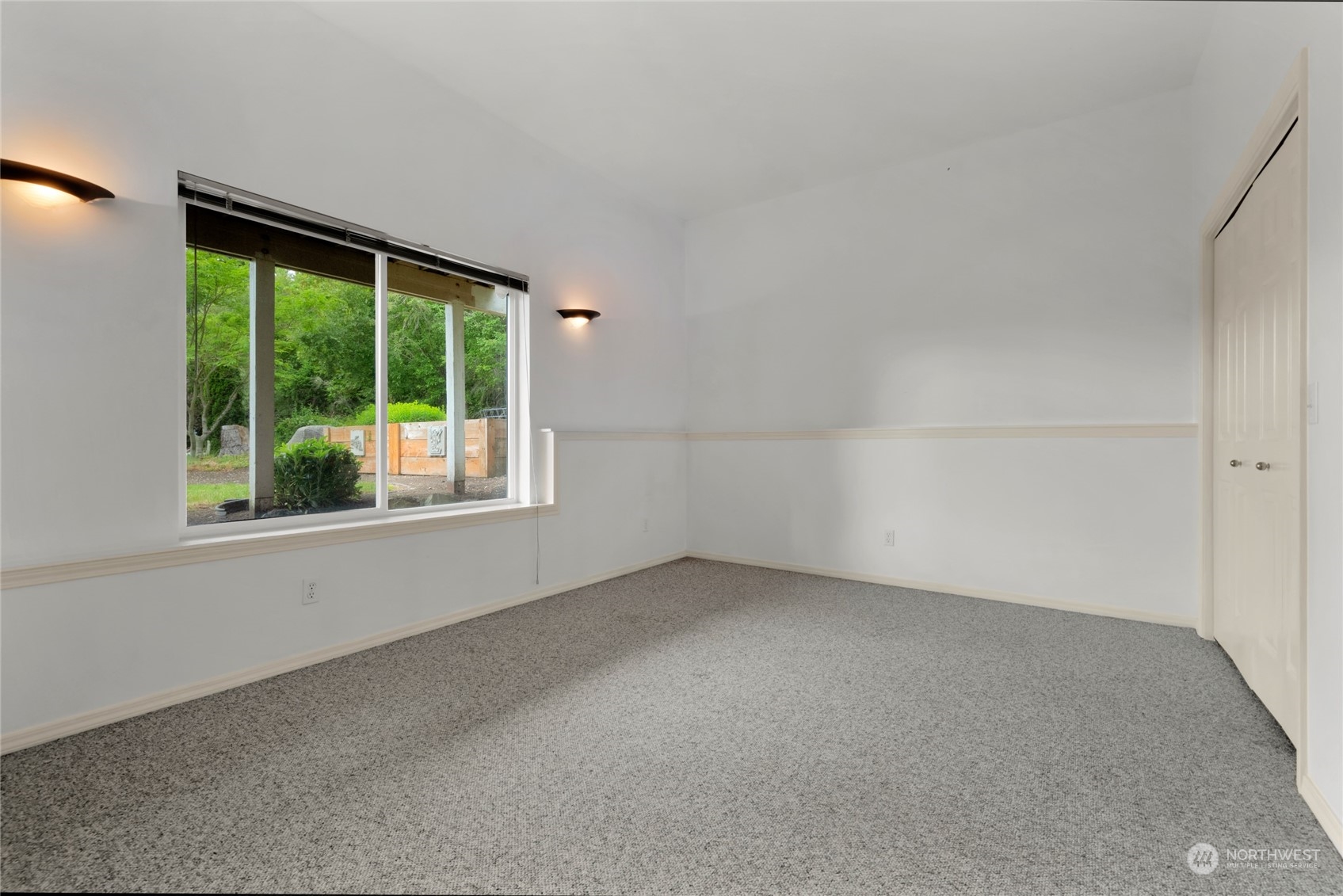 Empty carpeted room with a large window overlooking greenery and closet doors.