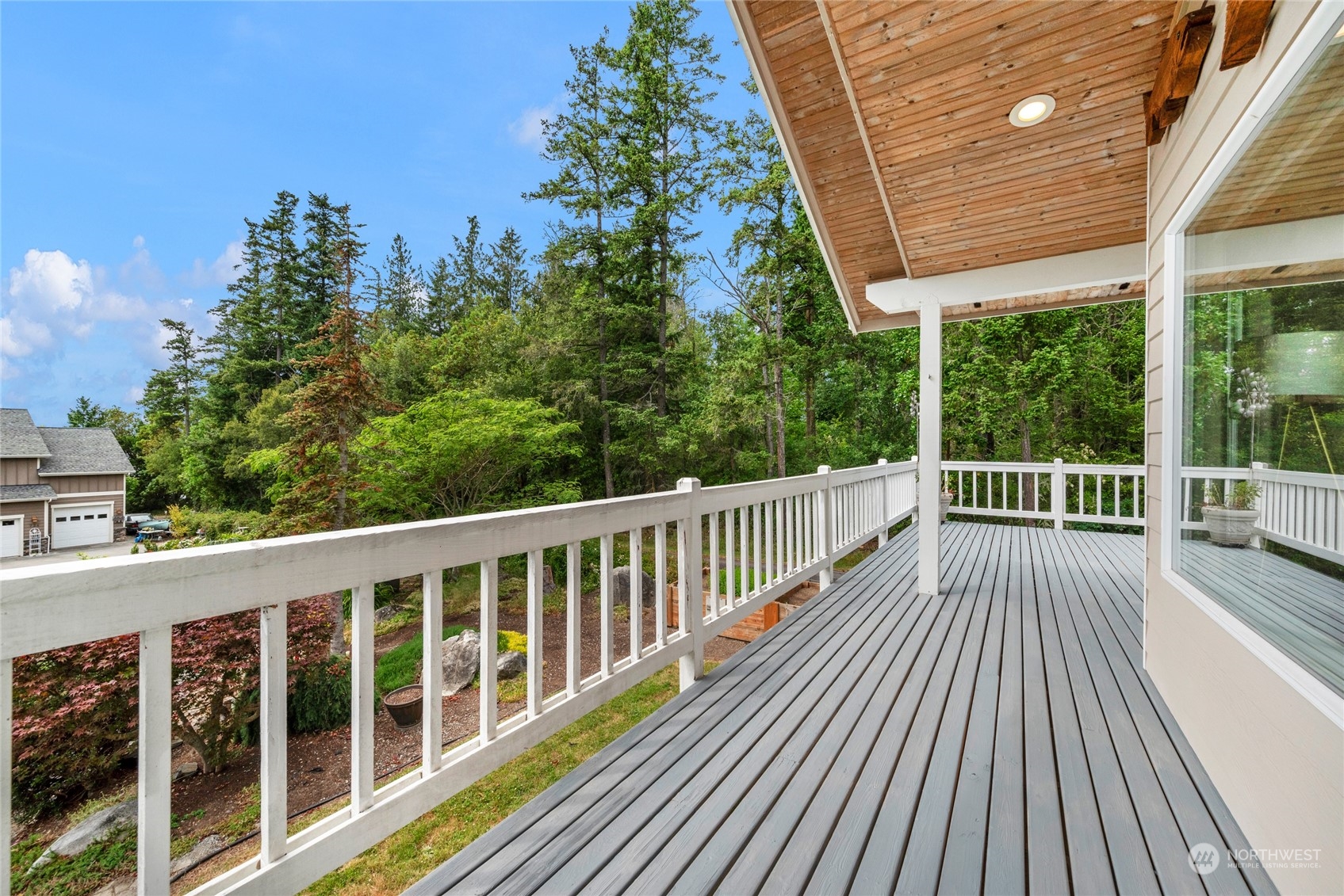 Covered deck with gray floorboards and white railings overlooking a leafy wooded yard.