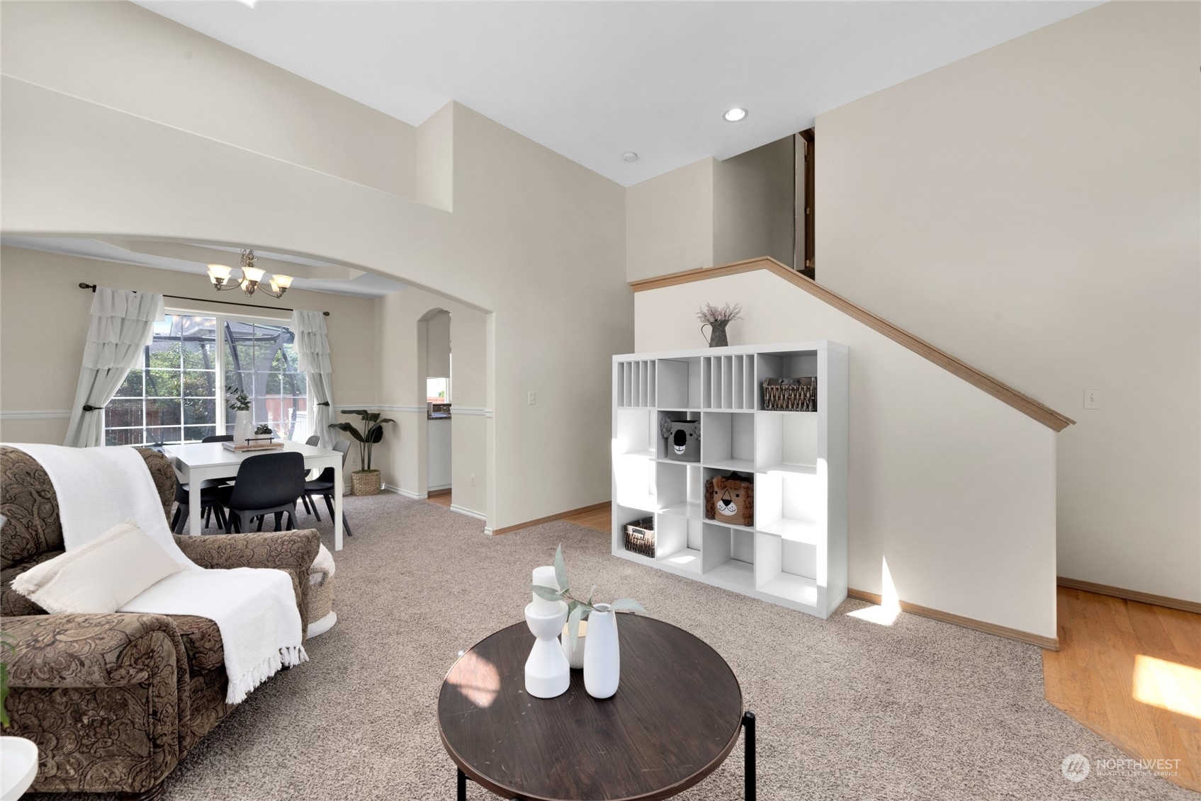 Living room with a carpeted floor, round coffee table, staircase, and dining area by a large window.