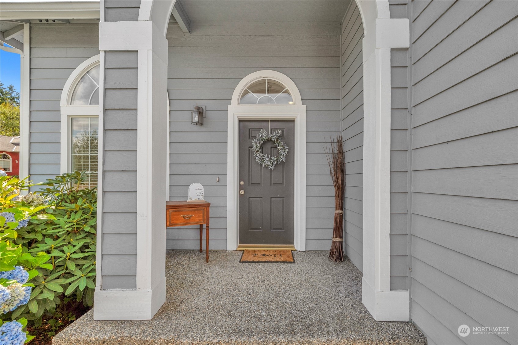 Covered front porch with gray siding, a dark door, arched window, and blue hydrangeas.