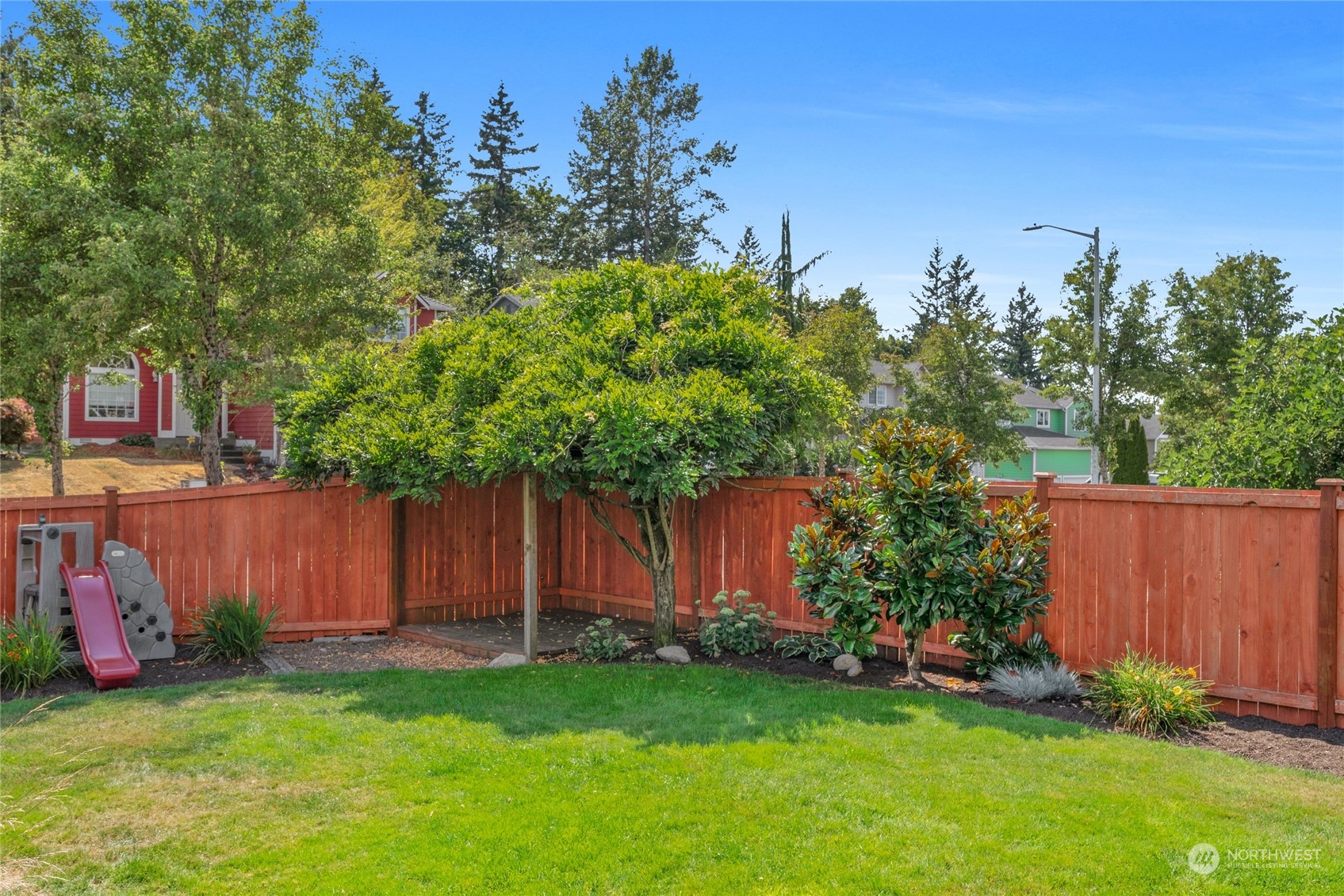 Backyard with a green lawn, red privacy fence, mature trees, landscaped shrubs, and a small play slide.