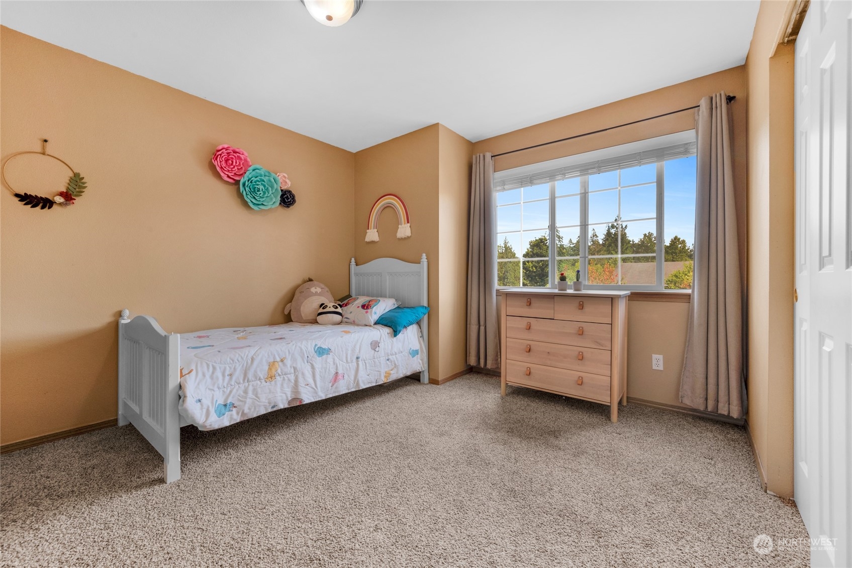 Bedroom with a white single bed, colorful wall decorations, and a large window overlooking trees.