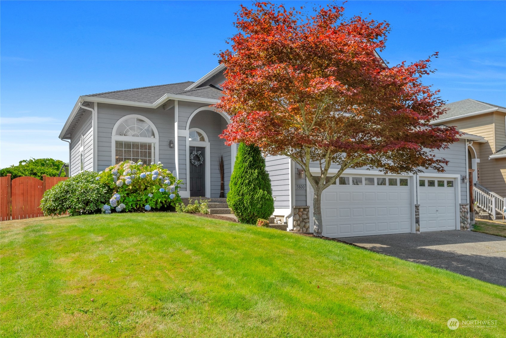 Gray home with an arched entry and white garage, framed by a large red-leaved tree.