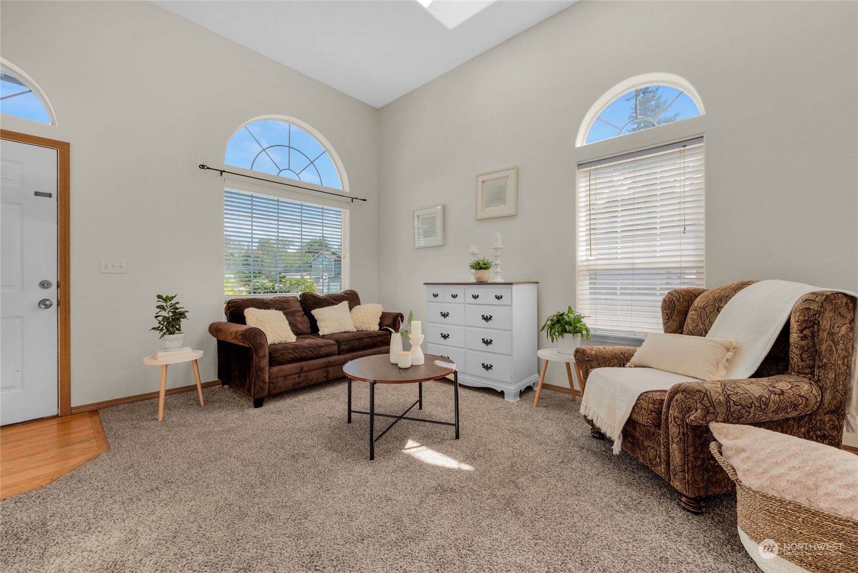 Staged living room with vaulted ceiling, arched windows, brown seating, and a white dresser.