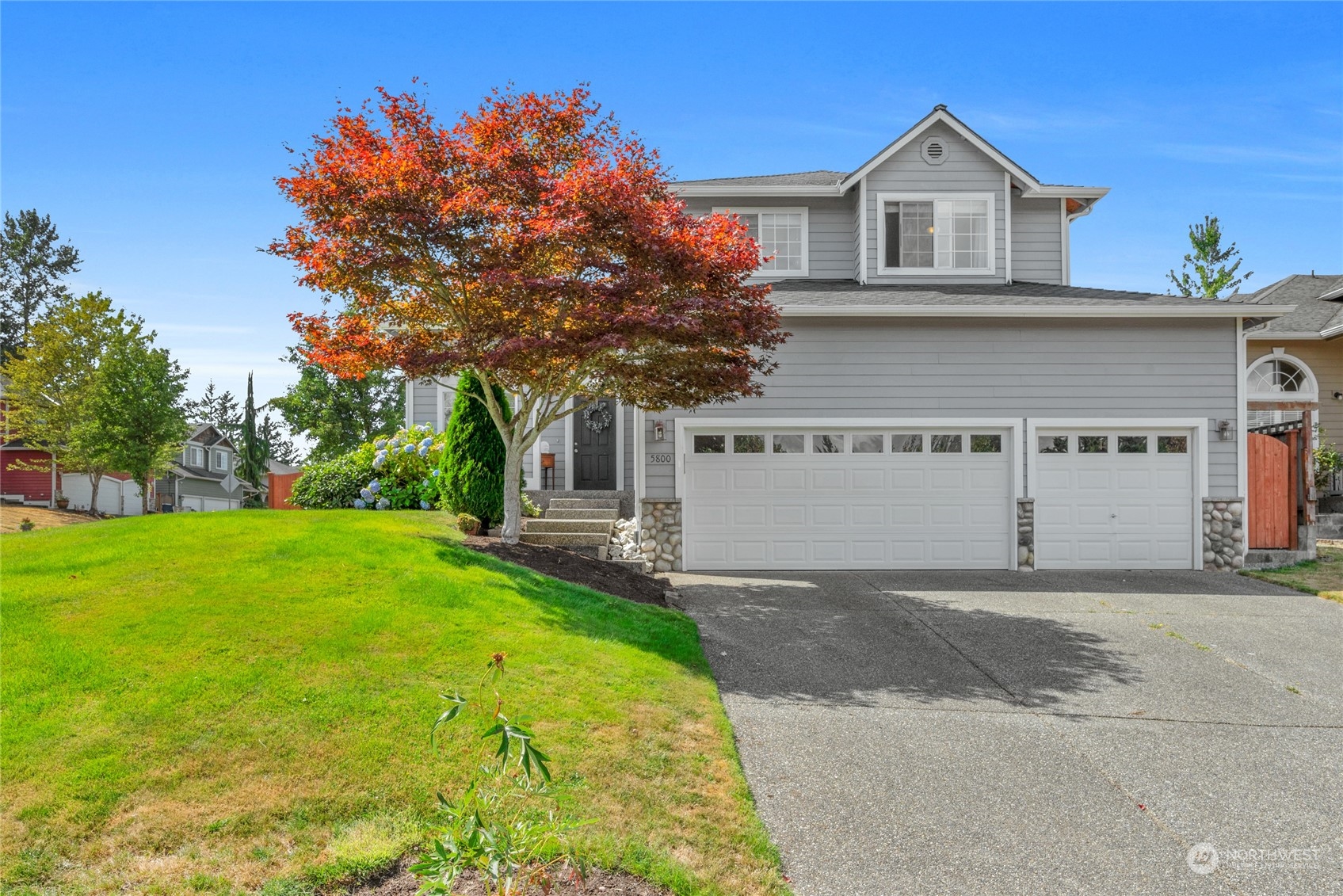 Gray two-story home with a broad driveway, three garage bays, and a red-orange autumn tree.