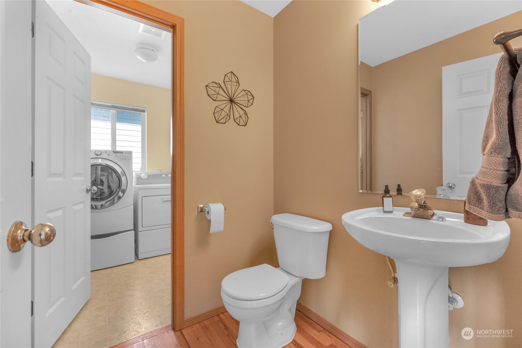 Tan bathroom with a pedestal sink and toilet; washer and dryer are visible through the open doorway.
