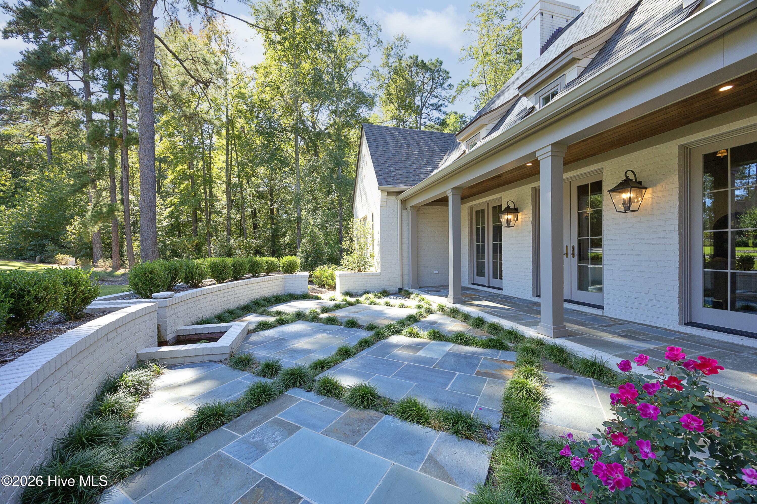 White brick home’s covered porch beside a slate courtyard with ornamental grasses and magenta flowers.