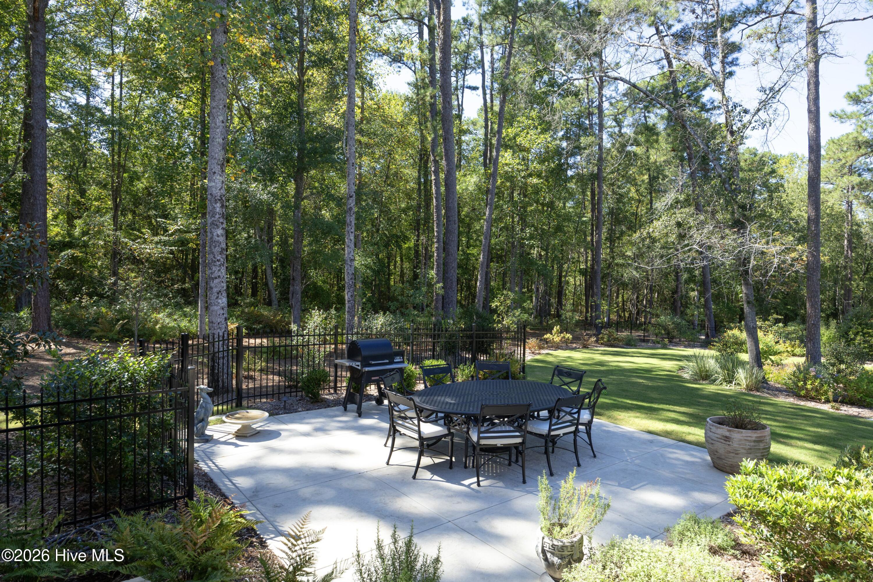 Outdoor dining patio with a metal table, grill, black fence, and wooded yard.