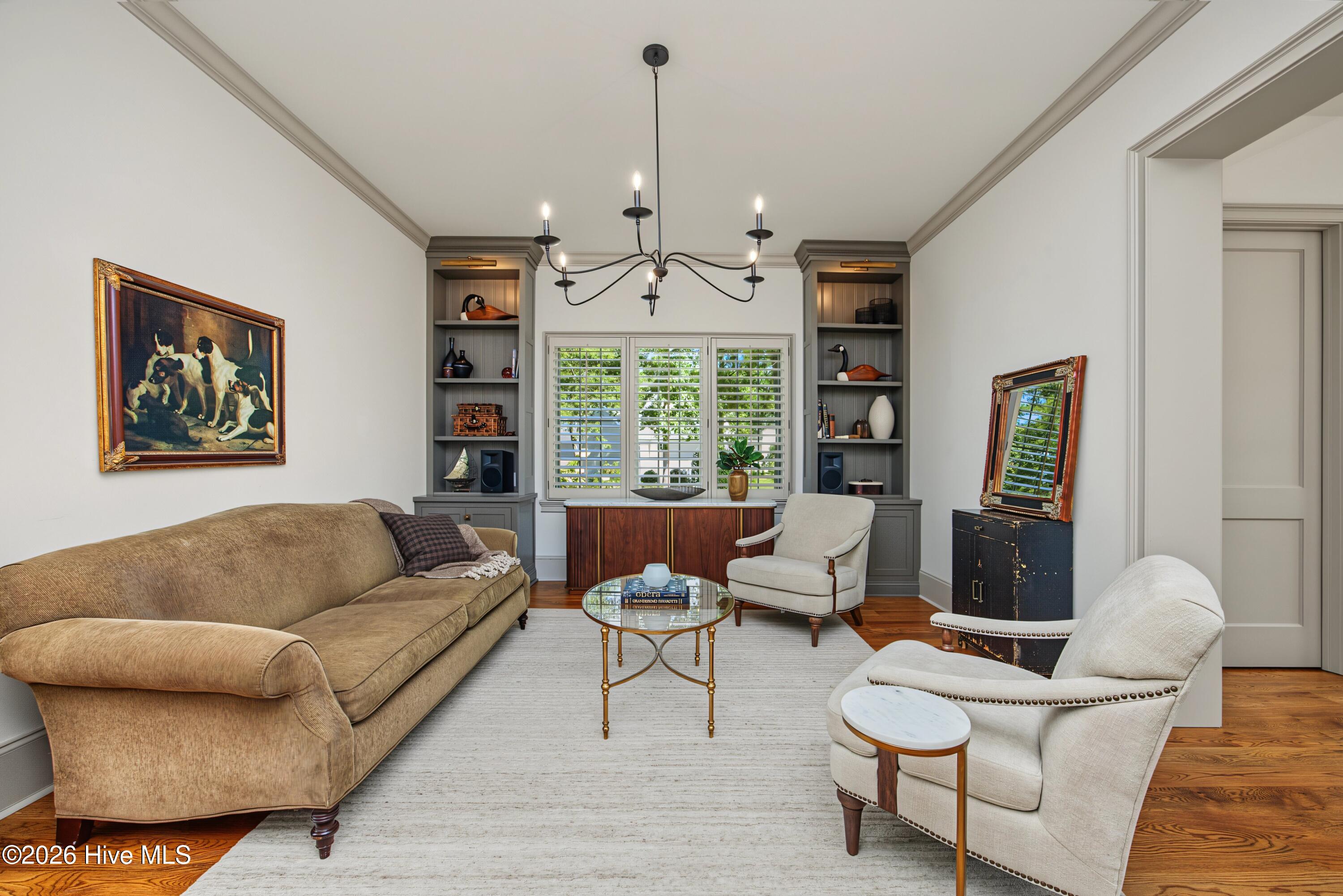Living room with a tan sofa, cream armchairs, built-in shelving, shuttered windows, and hardwood floors.