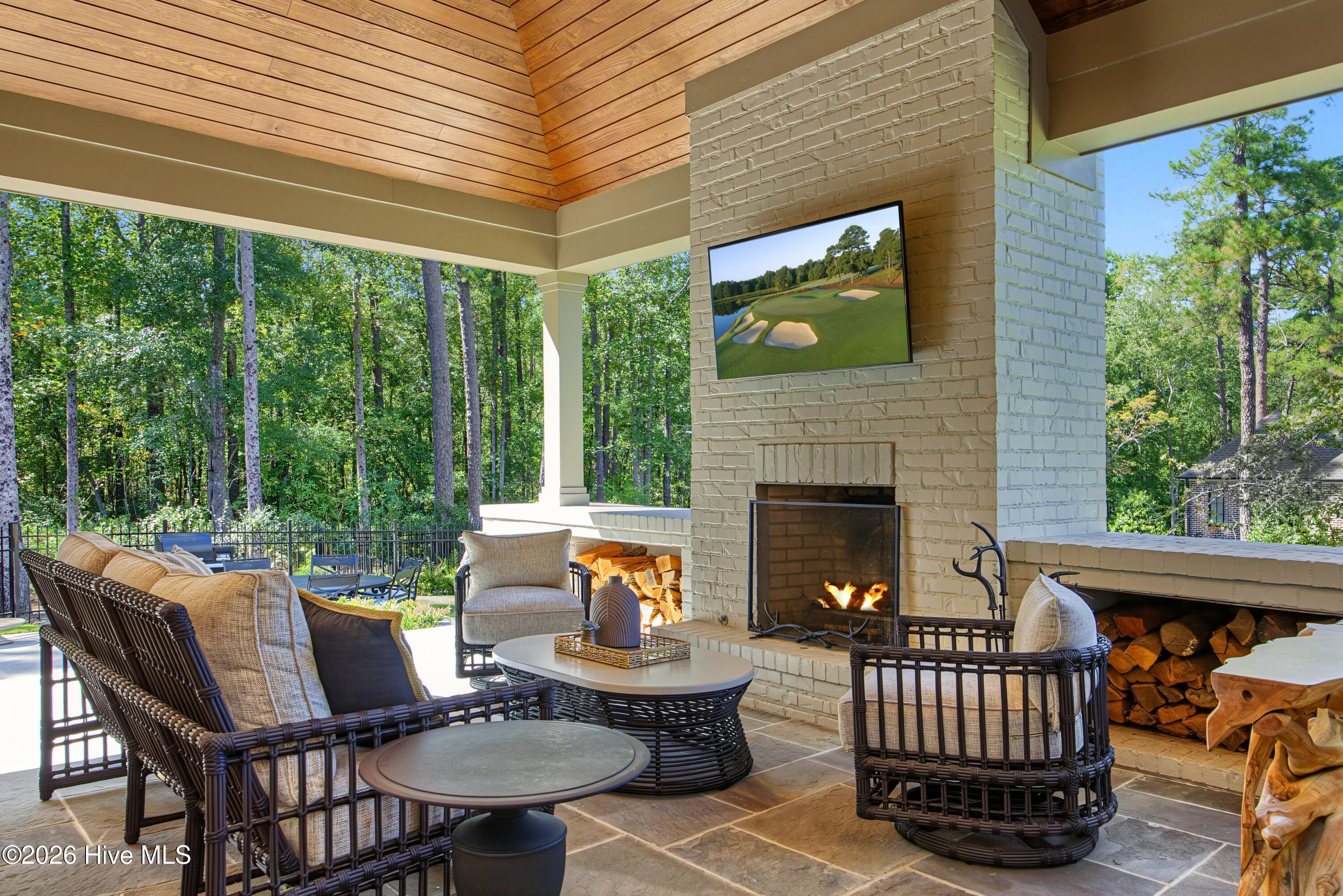 Covered patio with a vaulted wood ceiling, brick fireplace, and furnished seating overlooking trees.