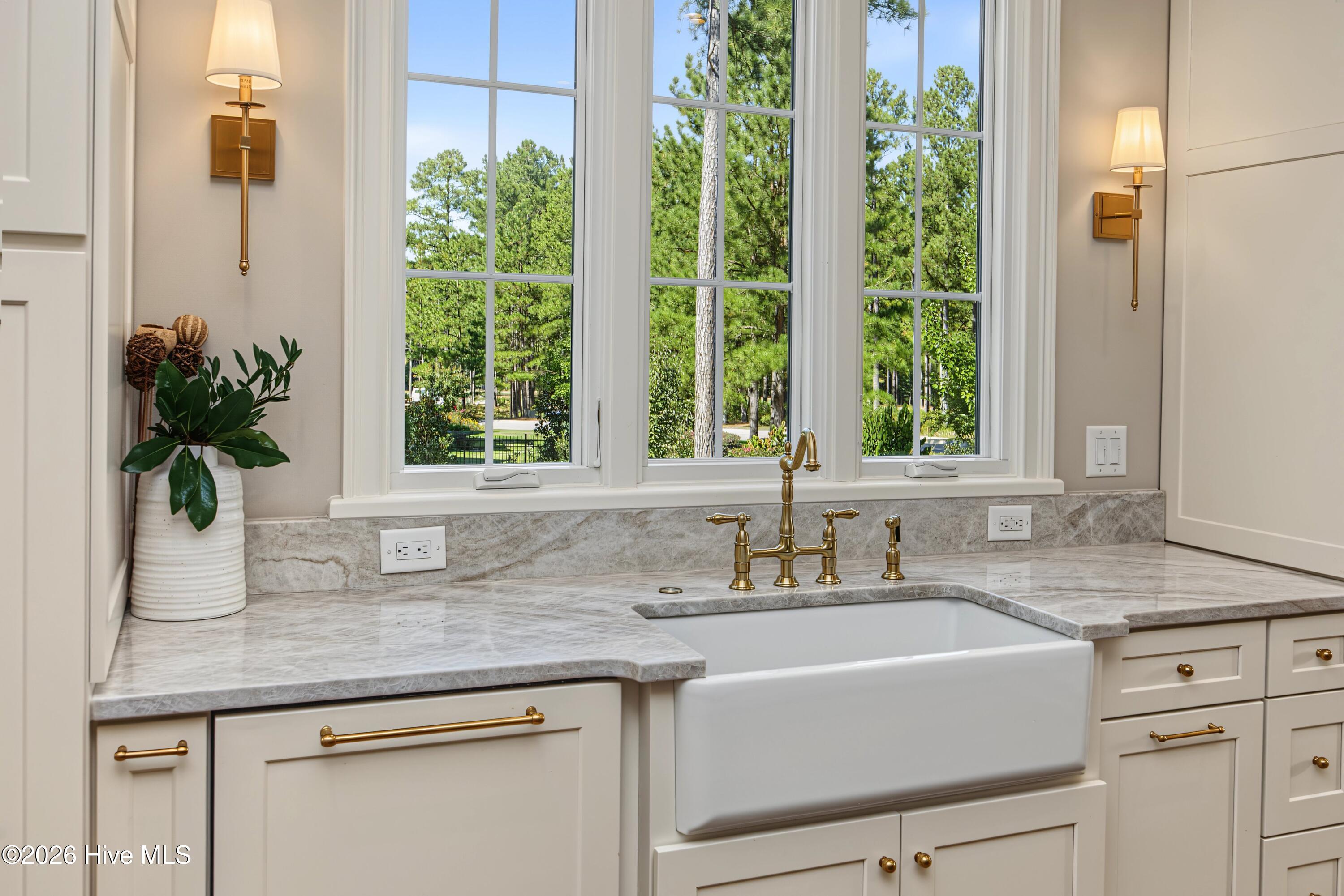 Kitchen sink with brass fixtures and marble counters beneath large windows overlooking trees.