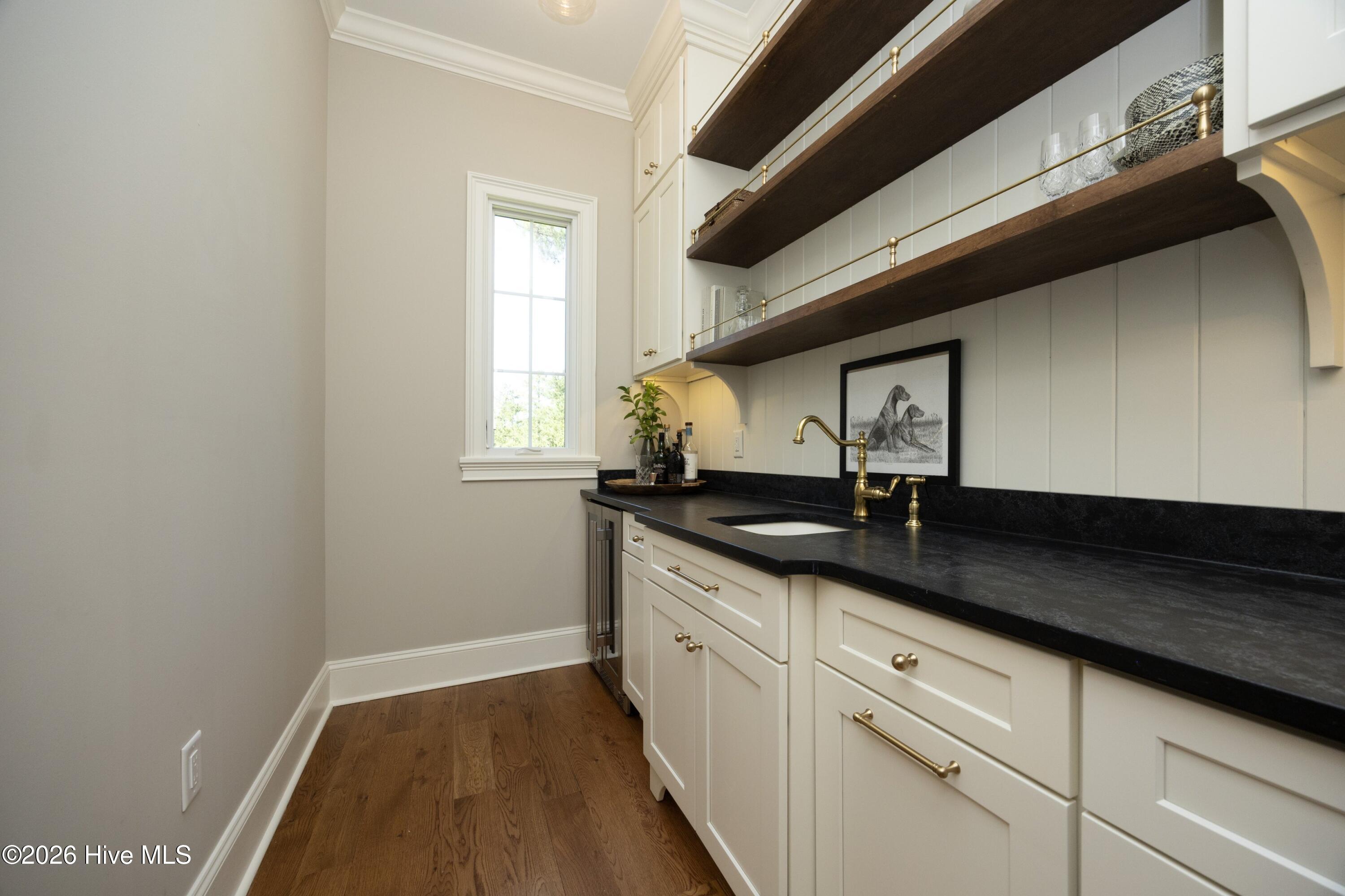 Wet bar with cream cabinetry, black counters, brass fixtures, and long dark wood shelves.