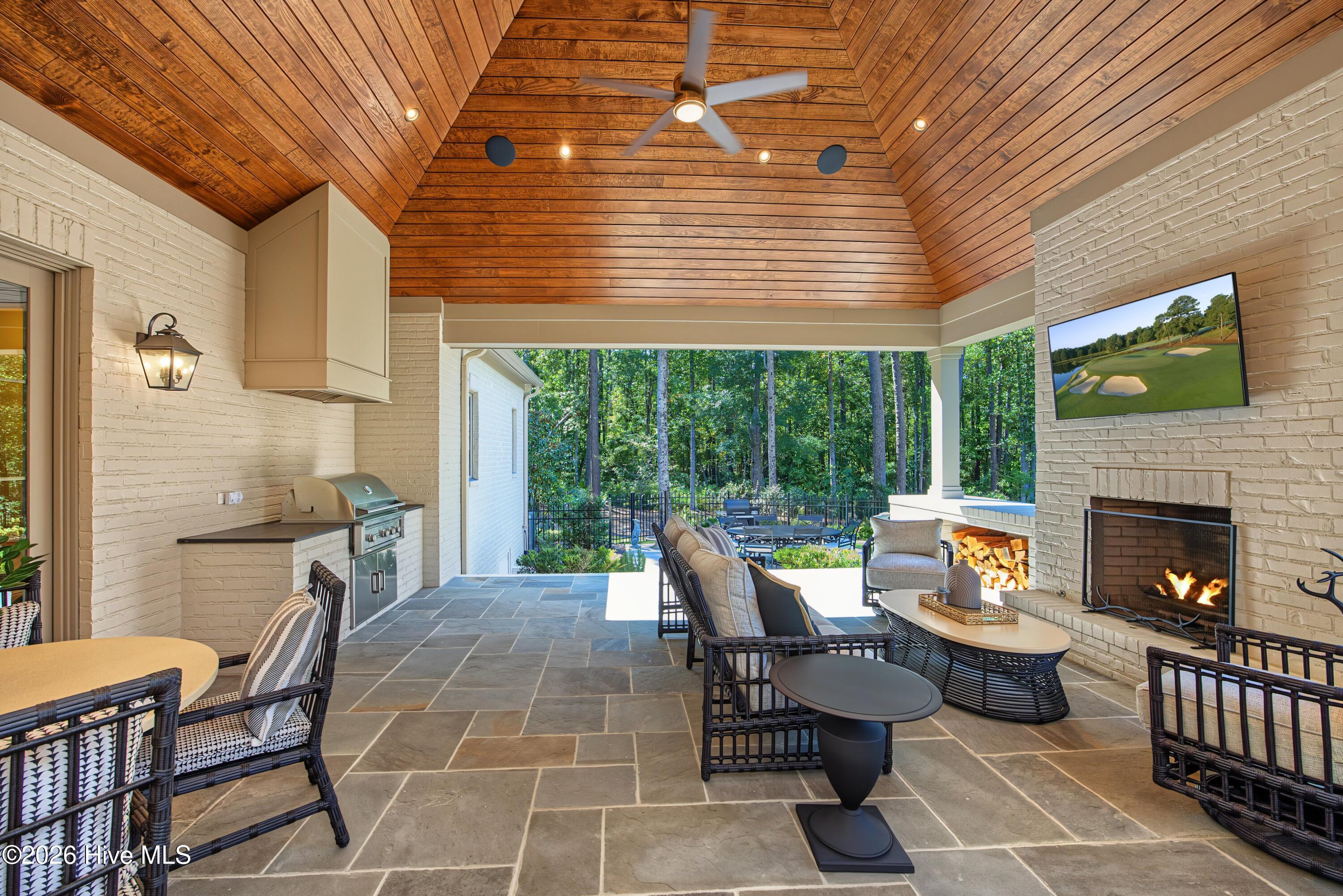 Covered patio with vaulted wood ceiling, fireplace, built-in grill, and seating overlooking the woods.