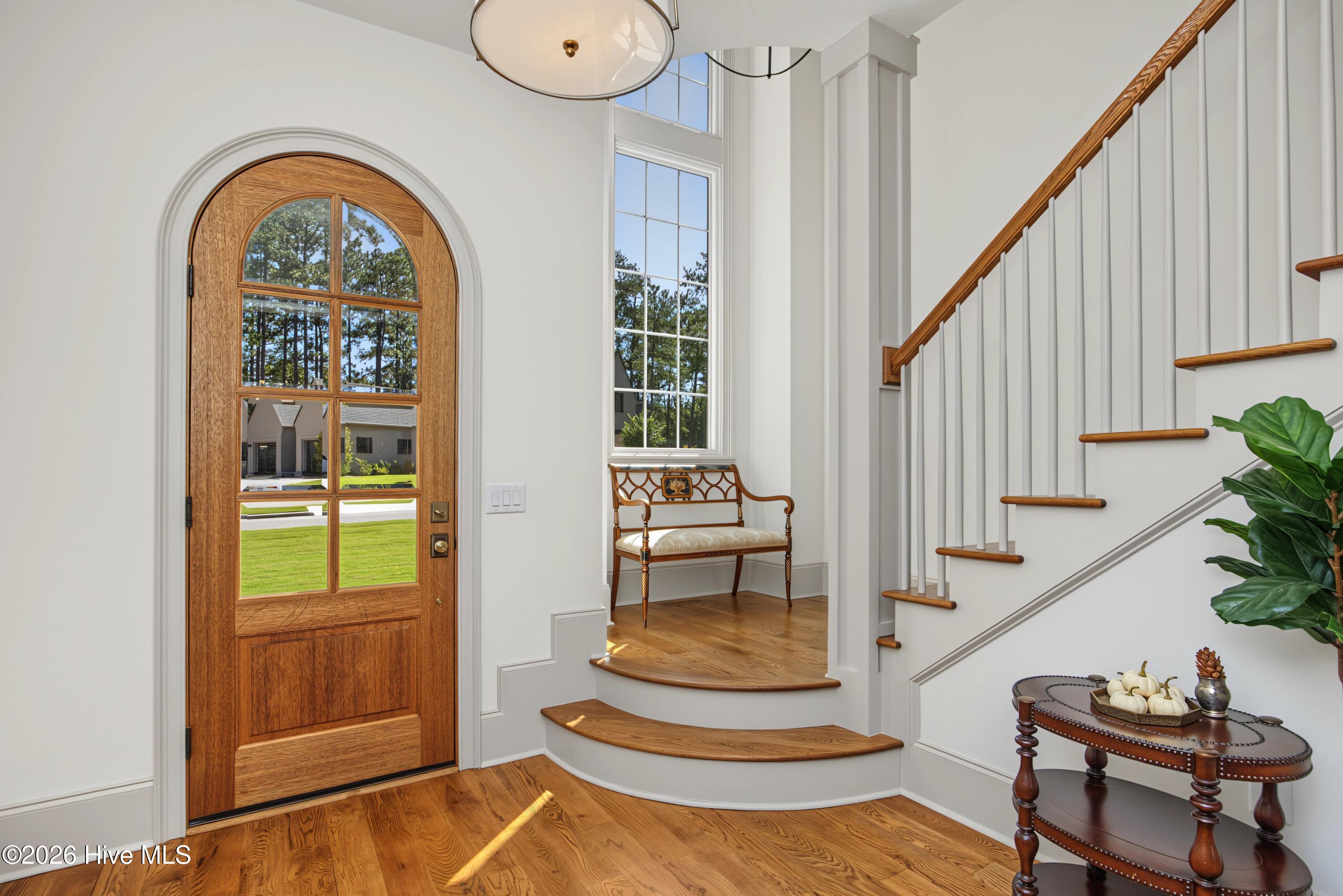 Foyer with an arched wood front door, curved staircase, hardwood floors, and a bench beneath a tall window.