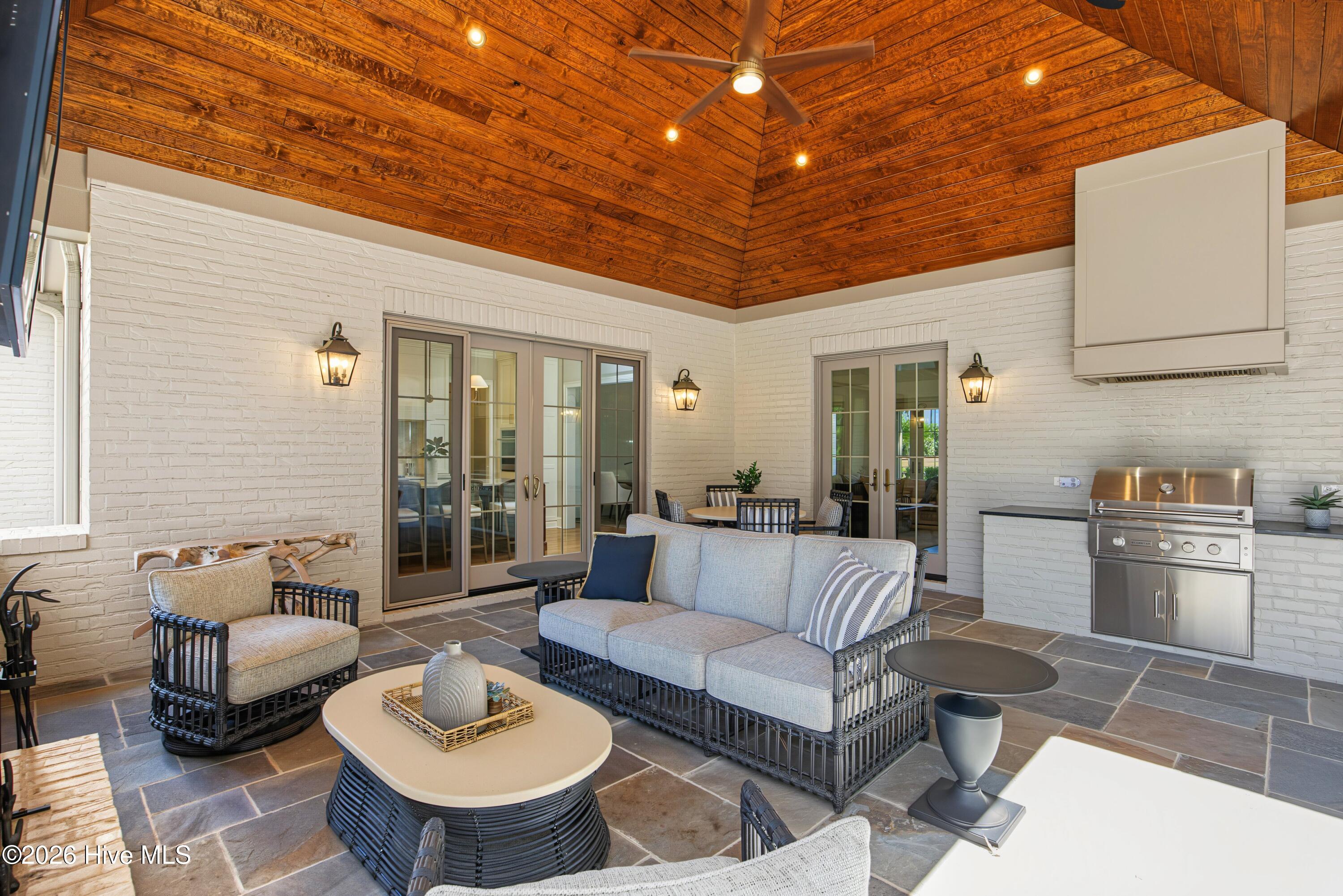 Covered patio with wicker seating, white brick walls, a wood-plank ceiling, and built-in stainless steel grill.
