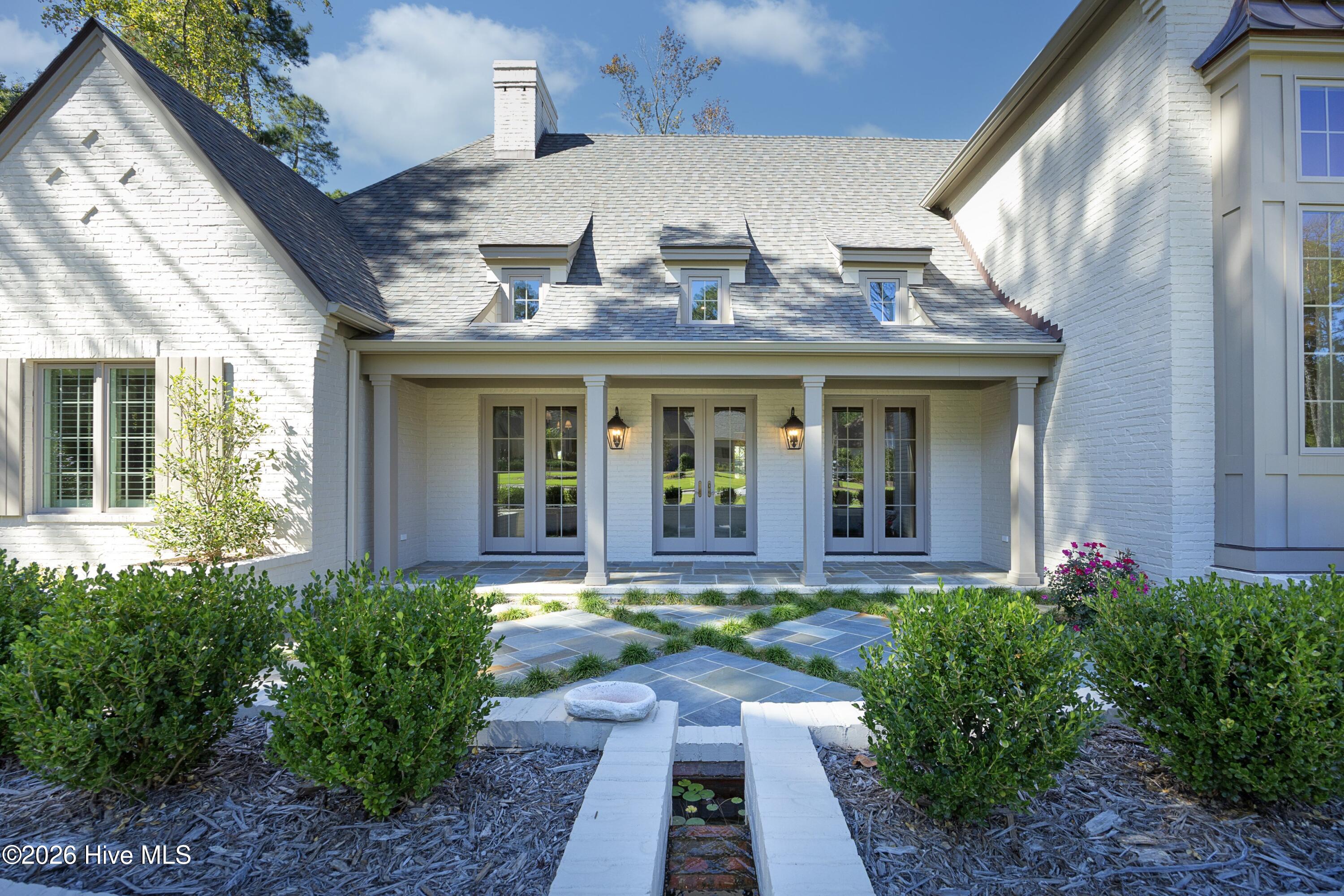 White brick home with a covered porch, stone patio, landscaped shrubs, and narrow water feature.