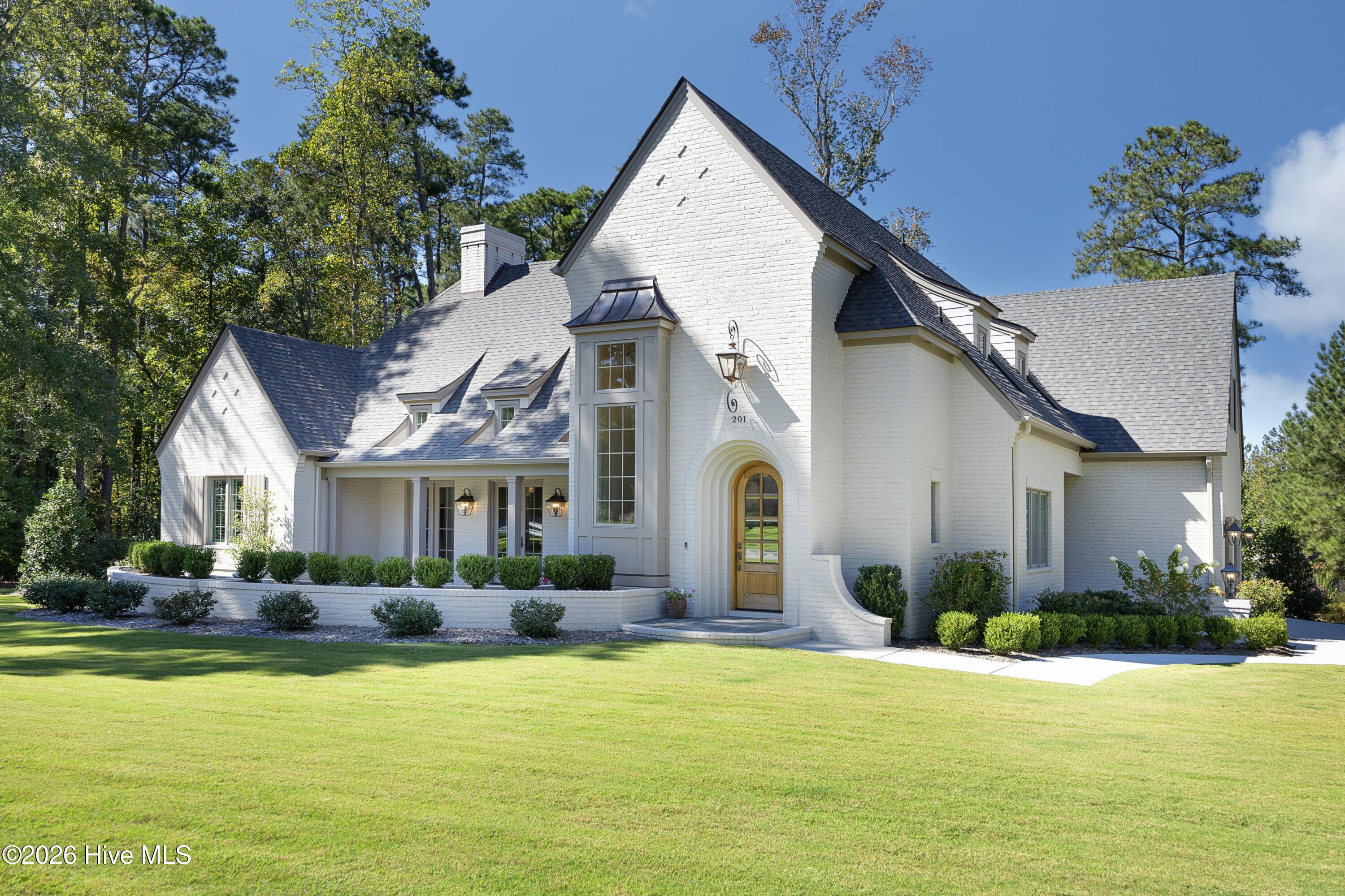 White brick house with steep gabled roofs, an arched wooden entry, landscaped shrubs, and a broad lawn.