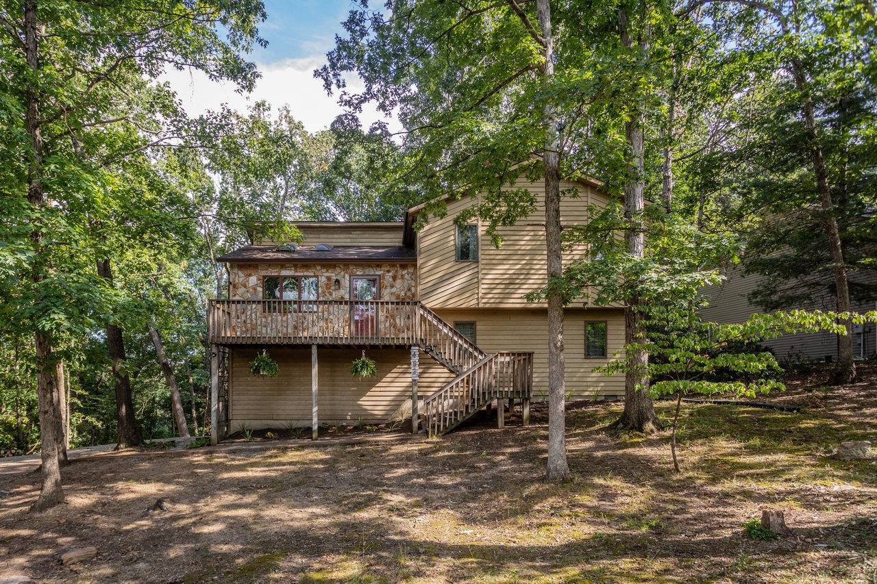 Two-story home with a stone-faced upper level and wooden deck, surrounded by mature trees.