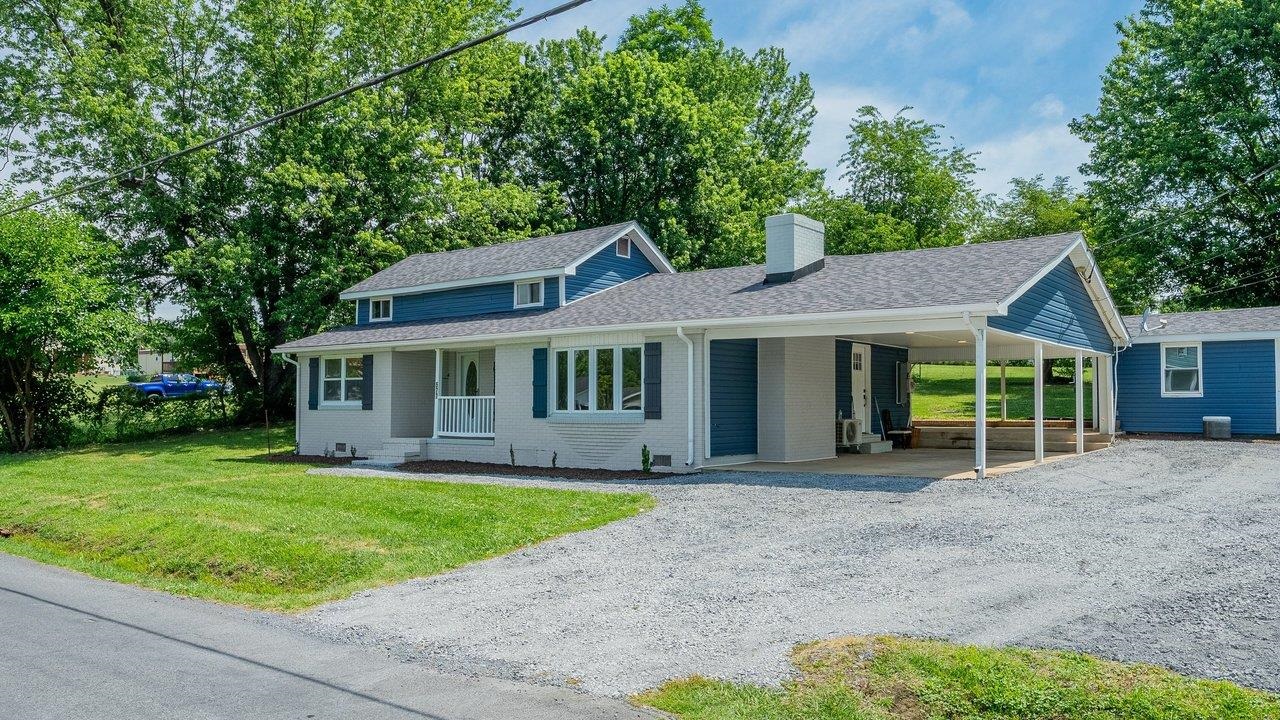 White brick and blue-sided home with a covered carport and gravel driveway beneath mature trees.