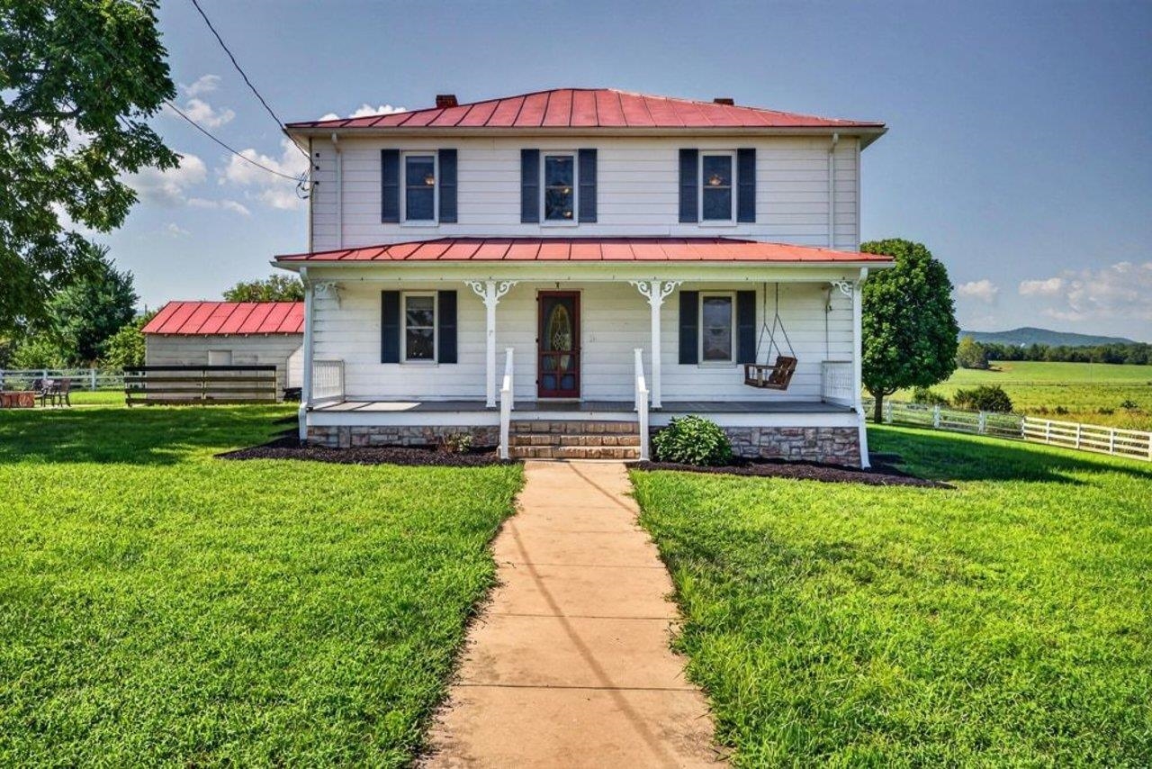 White two-story farmhouse with red metal roofs, a covered porch, and a walkway across its lawn.