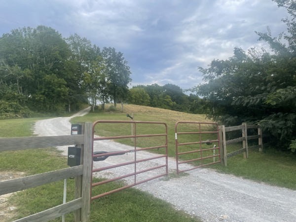 Gravel driveway through red metal gates, bordered by wooden fencing and a grassy field.