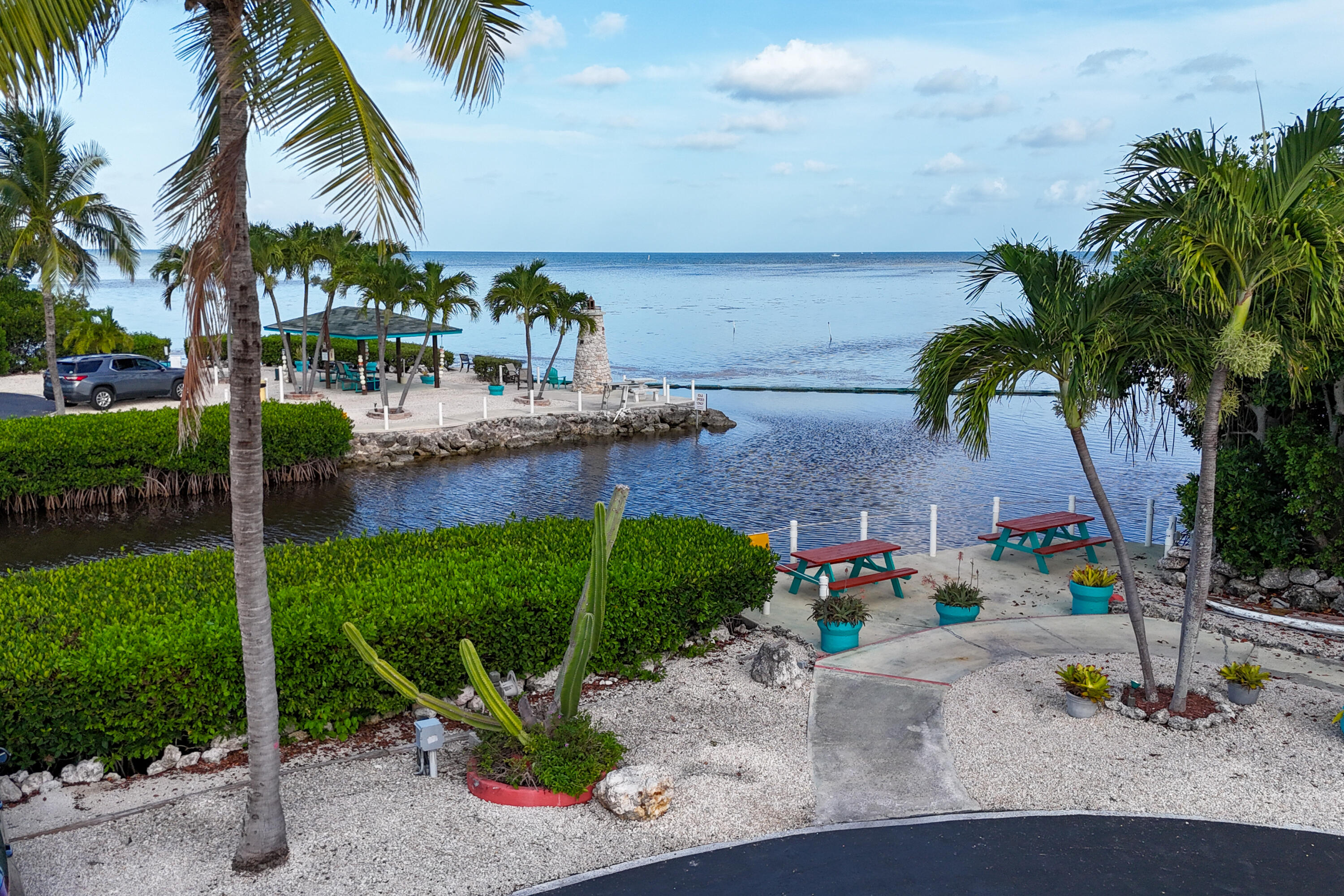 Waterfront picnic area with palm trees, turquoise tables, and a calm inlet opening to the sea.