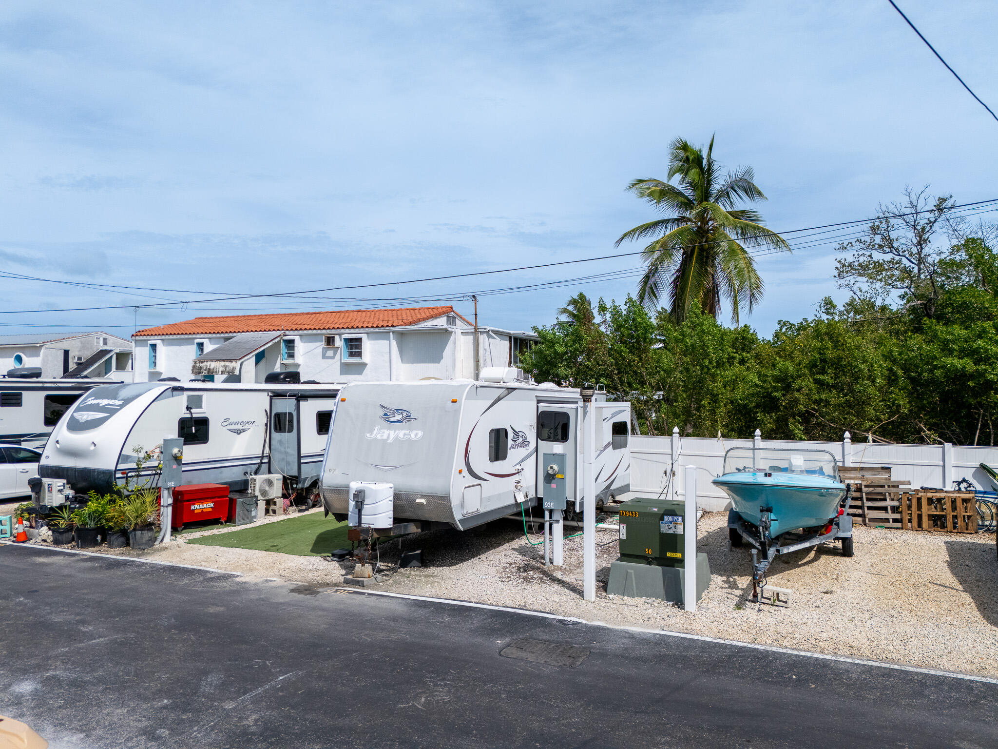 Property exterior with travel trailers and a blue boat beside a white fence and palm trees.