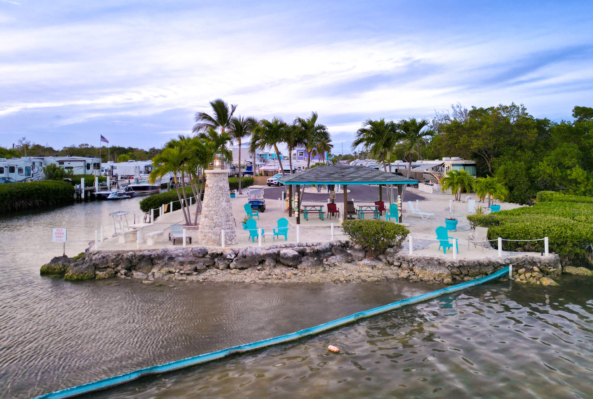 Waterfront pavilion with palm trees and turquoise chairs beside boats and a rocky shoreline.