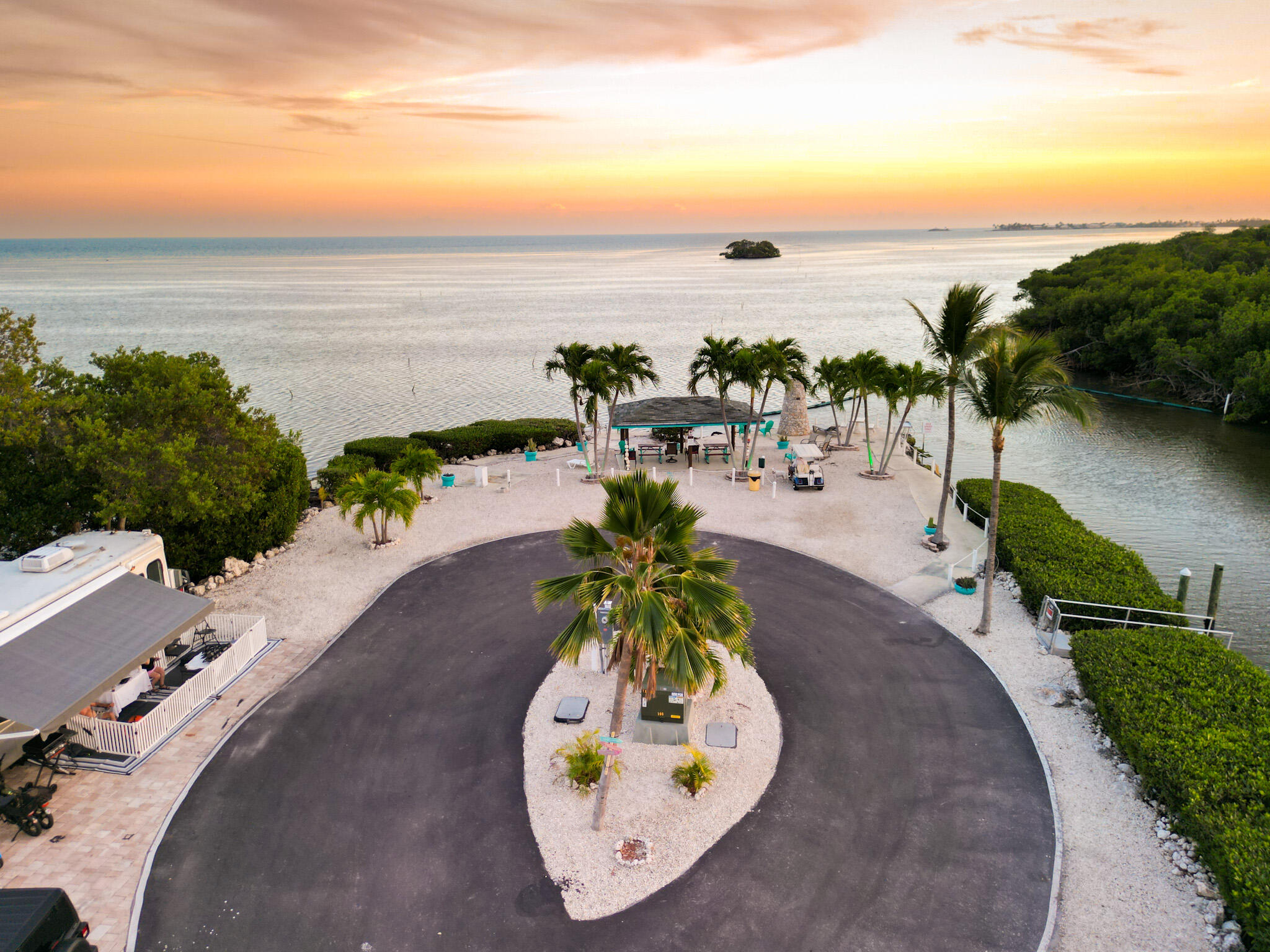 Waterfront property with a palm-lined drive, covered gathering area, and calm water at sunset.