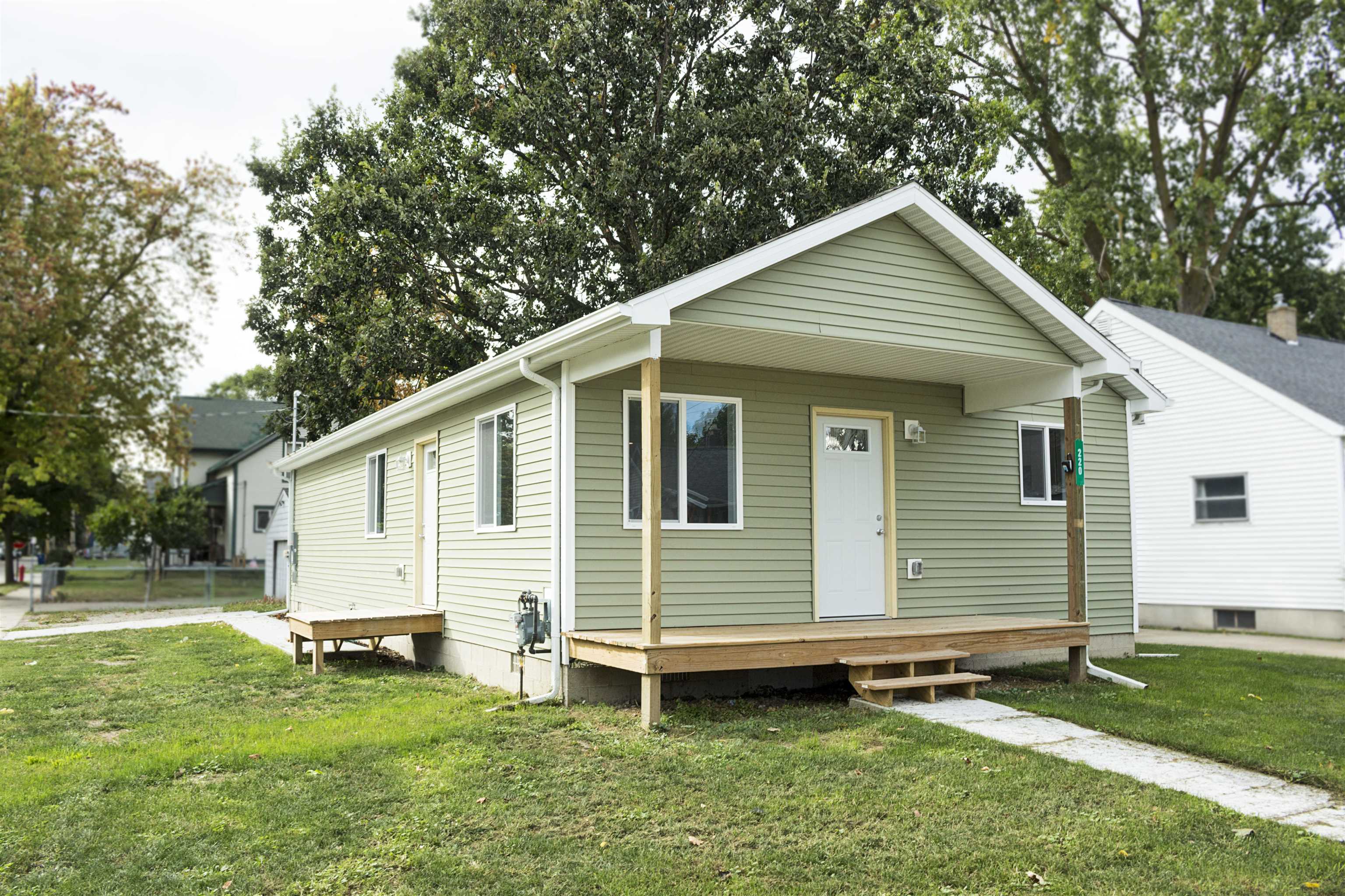 Single-story sage-green house with a covered porch, wooden steps, and a concrete walkway across the lawn.