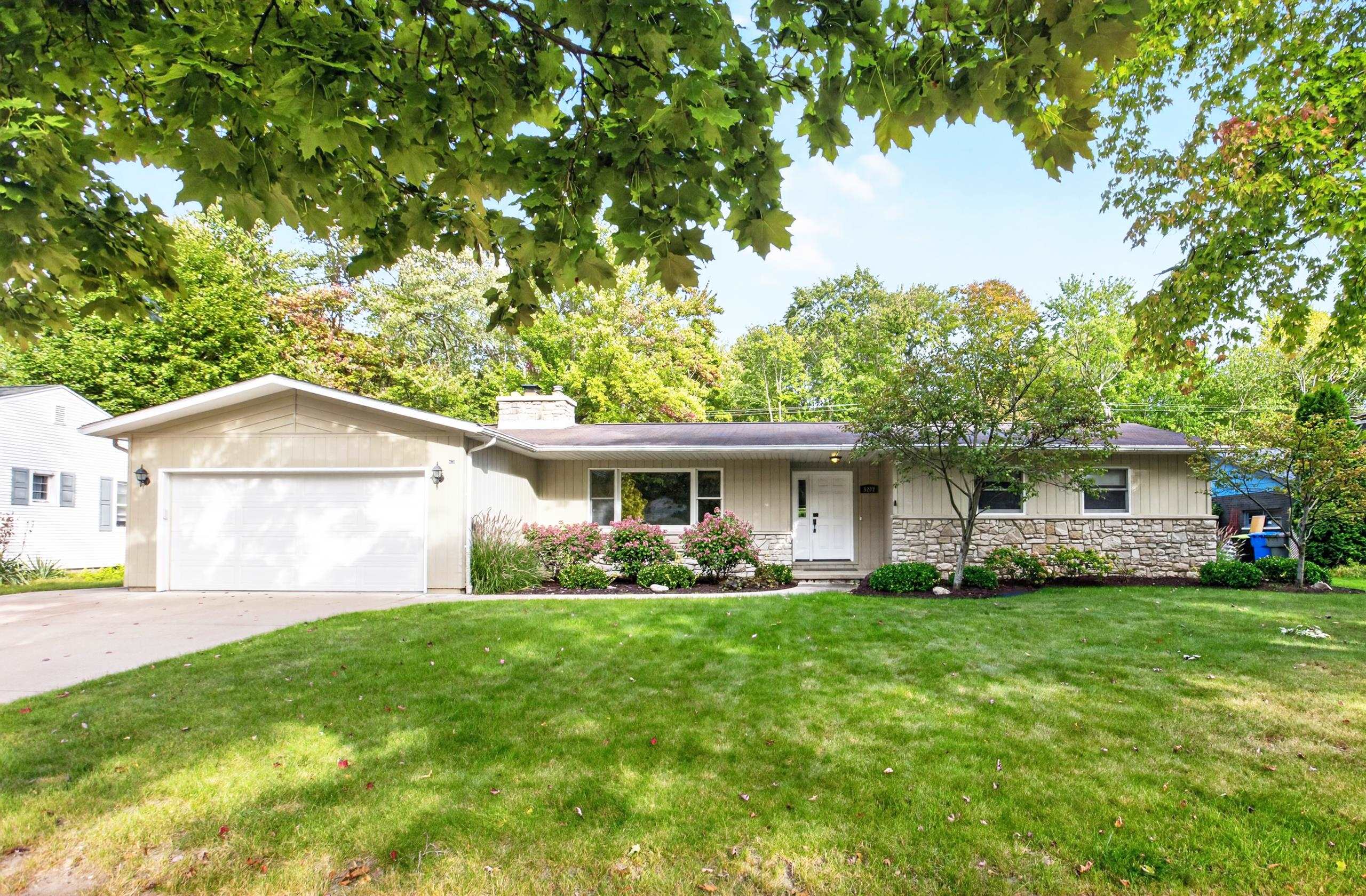 Single-story ranch home with stone façade, attached garage, landscaped shrubs, and a broad green lawn.