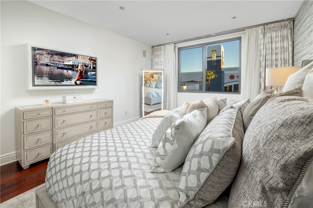 Furnished bedroom with a geometric-patterned bed, light wood dresser, and window overlooking nearby buildings.