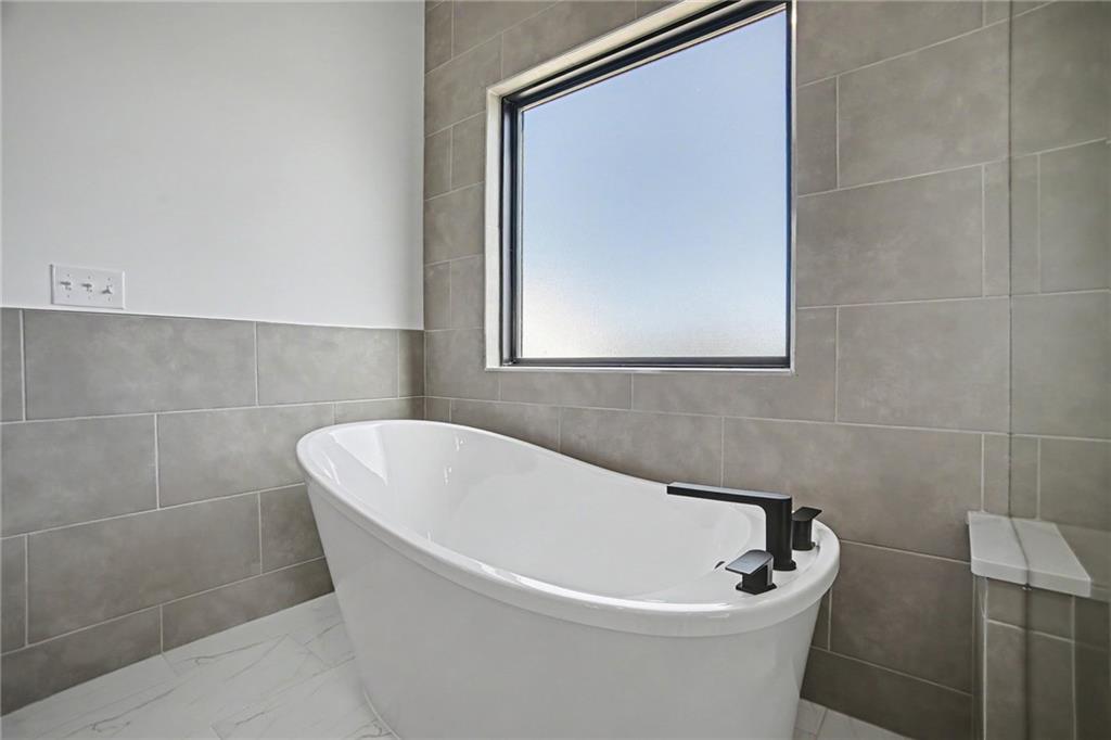 Modern bathroom with a white freestanding tub, black fixtures, gray tile, and a window.