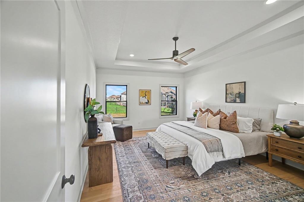Staged bedroom with a large white bed, patterned rug, ceiling fan, and two windows overlooking neighboring homes.