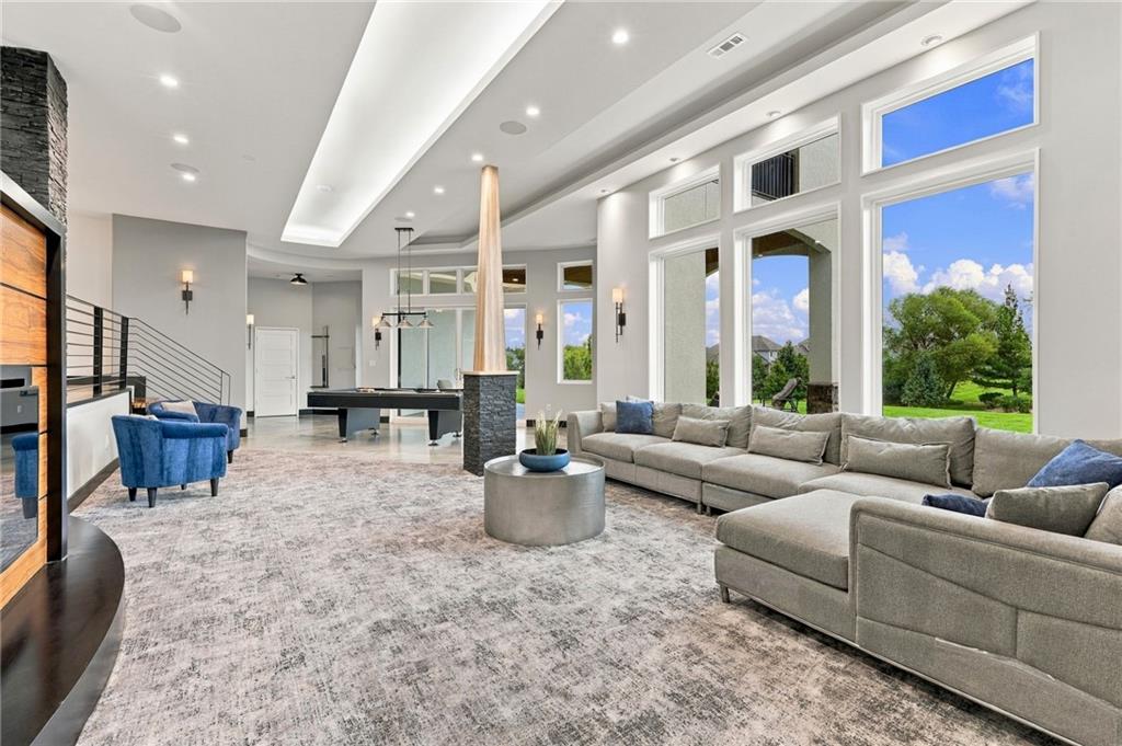 Modern living room with a gray sectional, blue chairs, tall windows, and an adjoining pool table.
