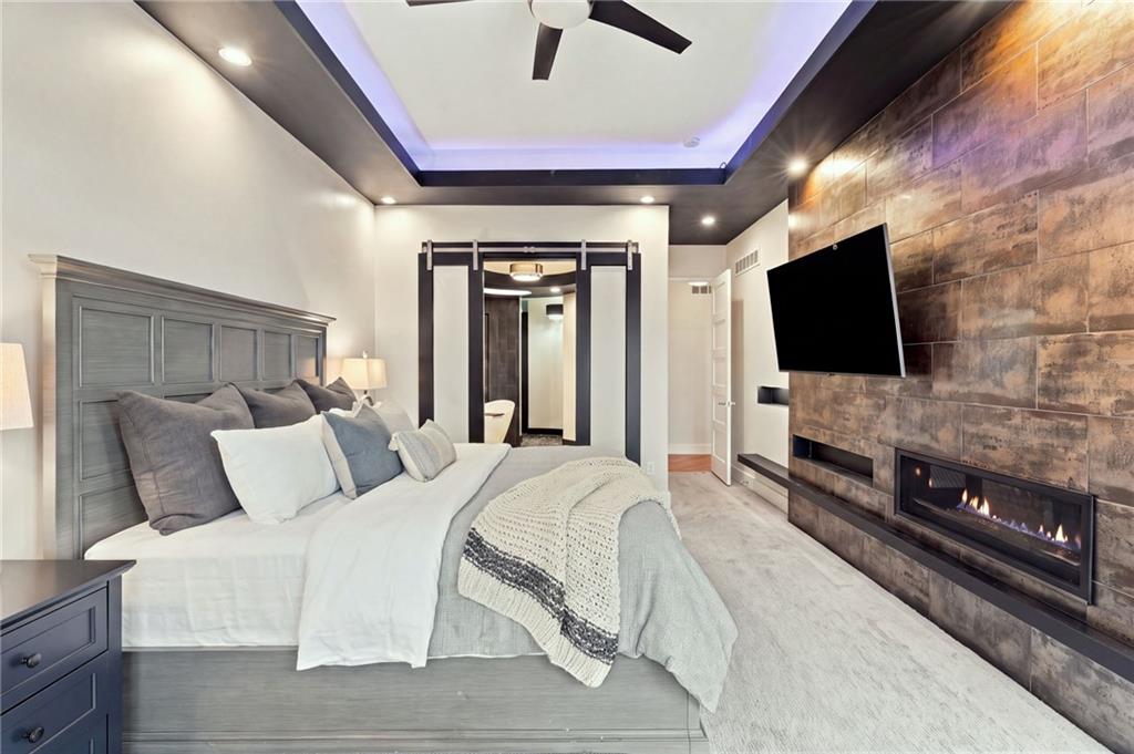 Bedroom with gray paneled bed, tray ceiling, and a television above a linear fireplace on a stone-tile wall.