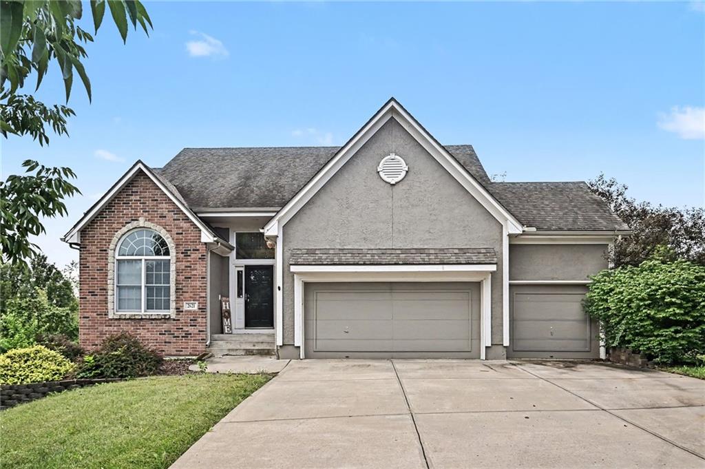 Gray stucco and brick house with a three-car garage, arched window, and broad concrete driveway.
